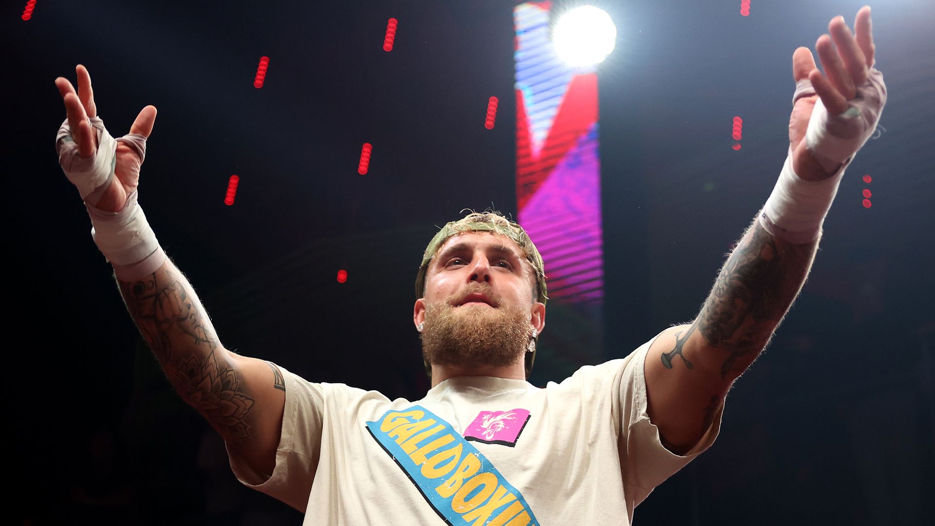 Bearded man with tattoos, wearing a beige "Gallo Boxing" t-shirt and a backwards cap, raising hands wrapped in white tape in a dark setting with bright light and red dots in background.