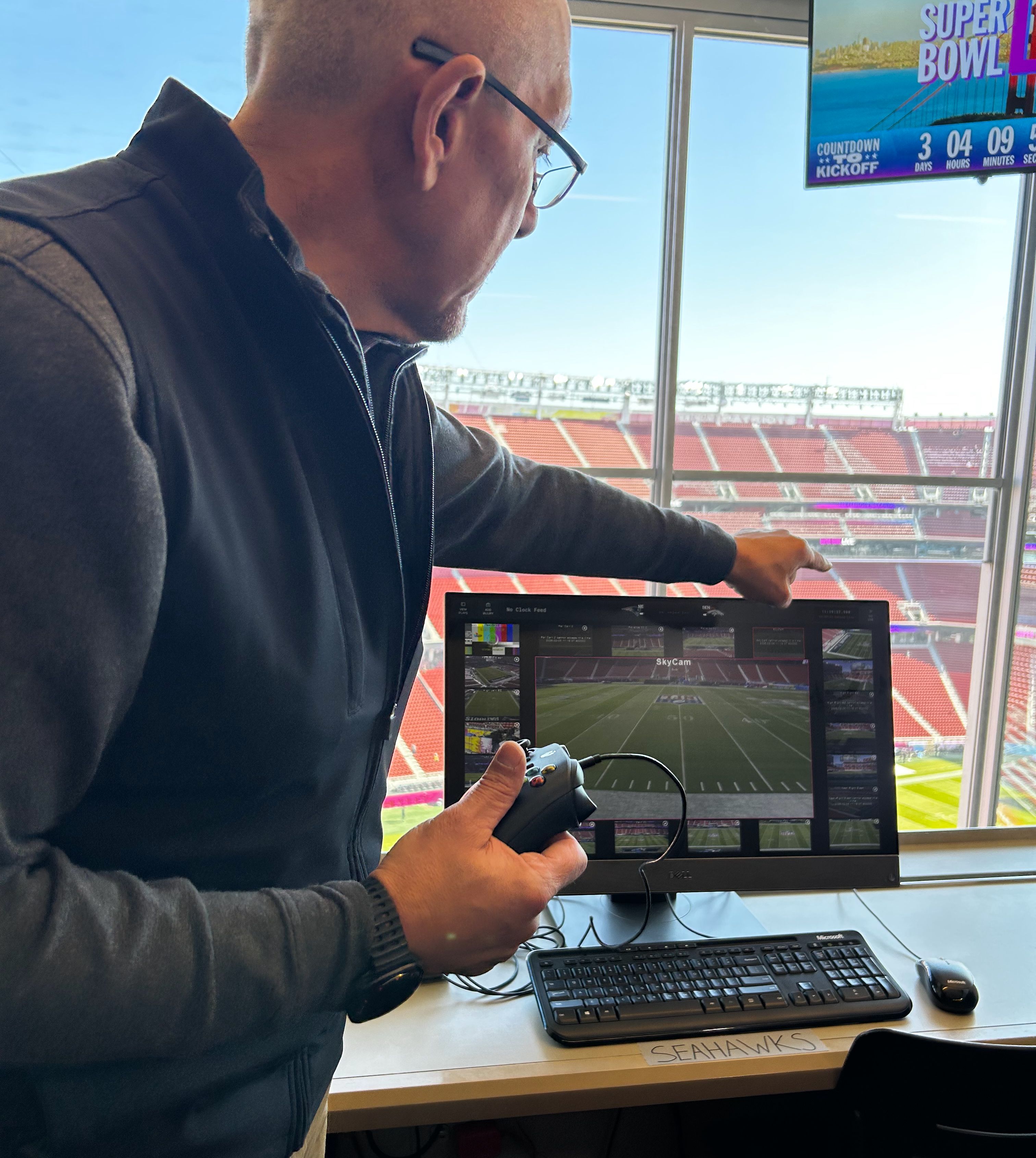 Man holding a game controller points at a computer screen showing a football field labeled SkyCam, inside a stadium with red seats and a Super Bowl countdown on a TV above.