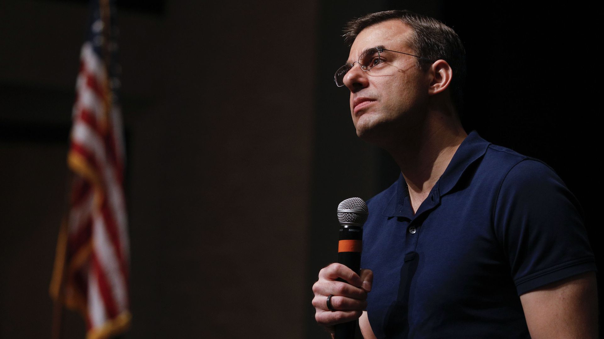 Justin Amash staring into the crowd at a town hall while holding a microphone. There is an American flag in the background and he is wearing a blue polo shirt and glasses. 