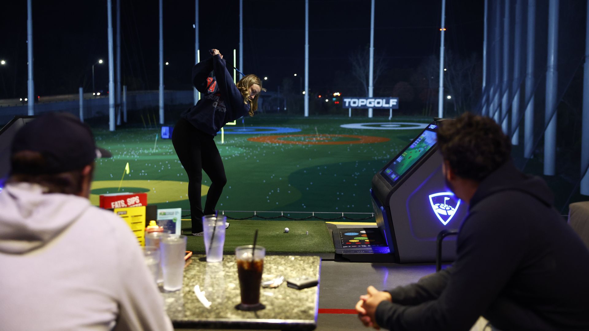 Night scene at a Topgolf driving range with a person swinging a golf club, two people watching, and a table with drinks and menus in the foreground.