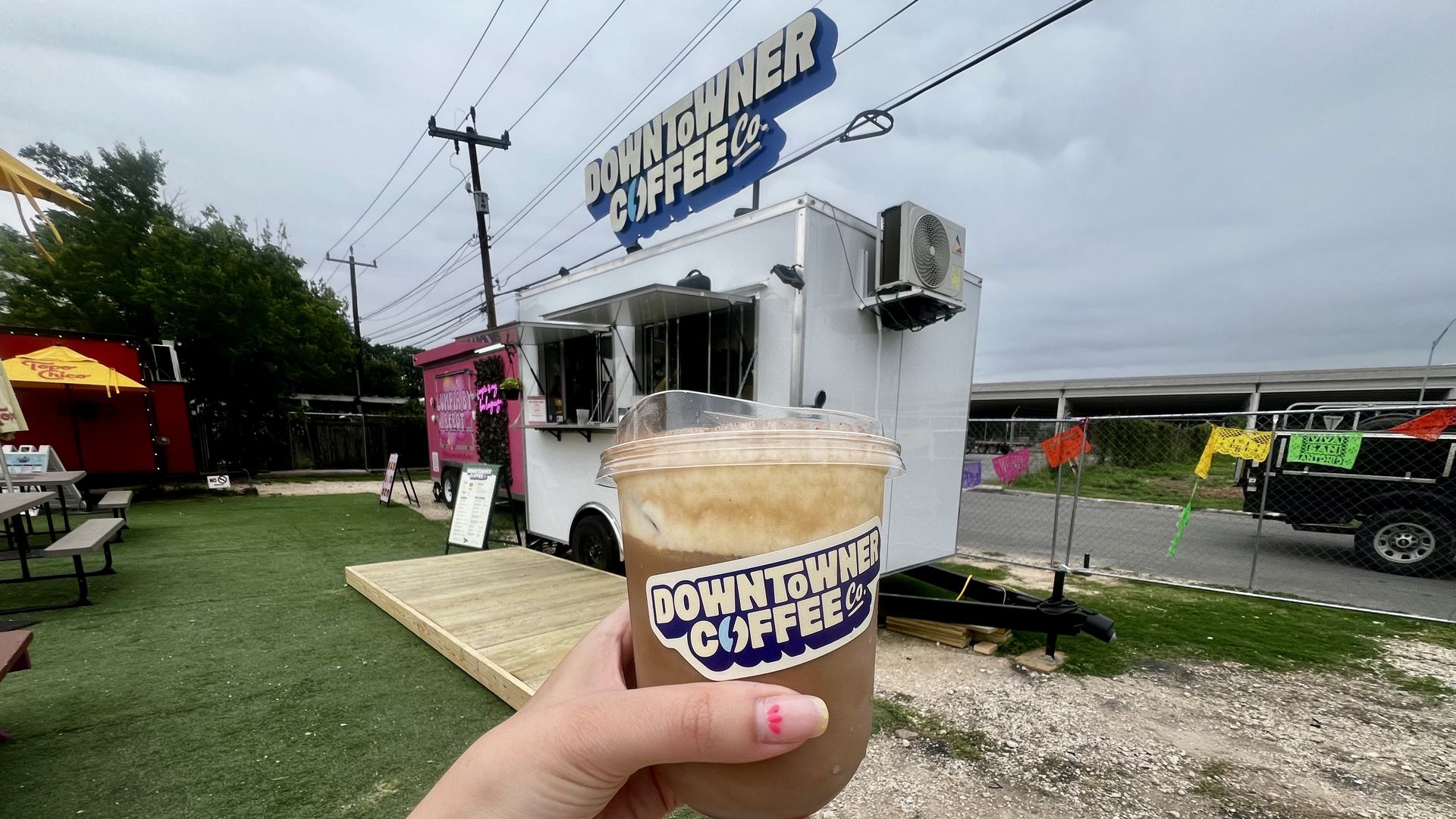 A hand holds a large iced coffee cup labeled "Downtown Coffee Co." in front of a white food truck outside Tony’s Siesta in San Antonio.