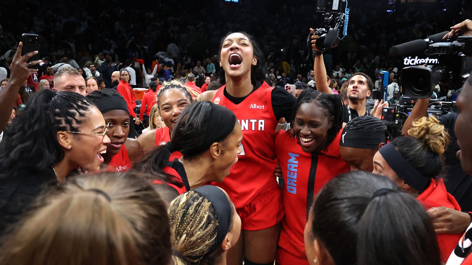 A women's basketball team wearing red "ATLANTA" jerseys celebrate a victory in an arena. Photographers circle the huddled players, one of whom is jumping and yelling.