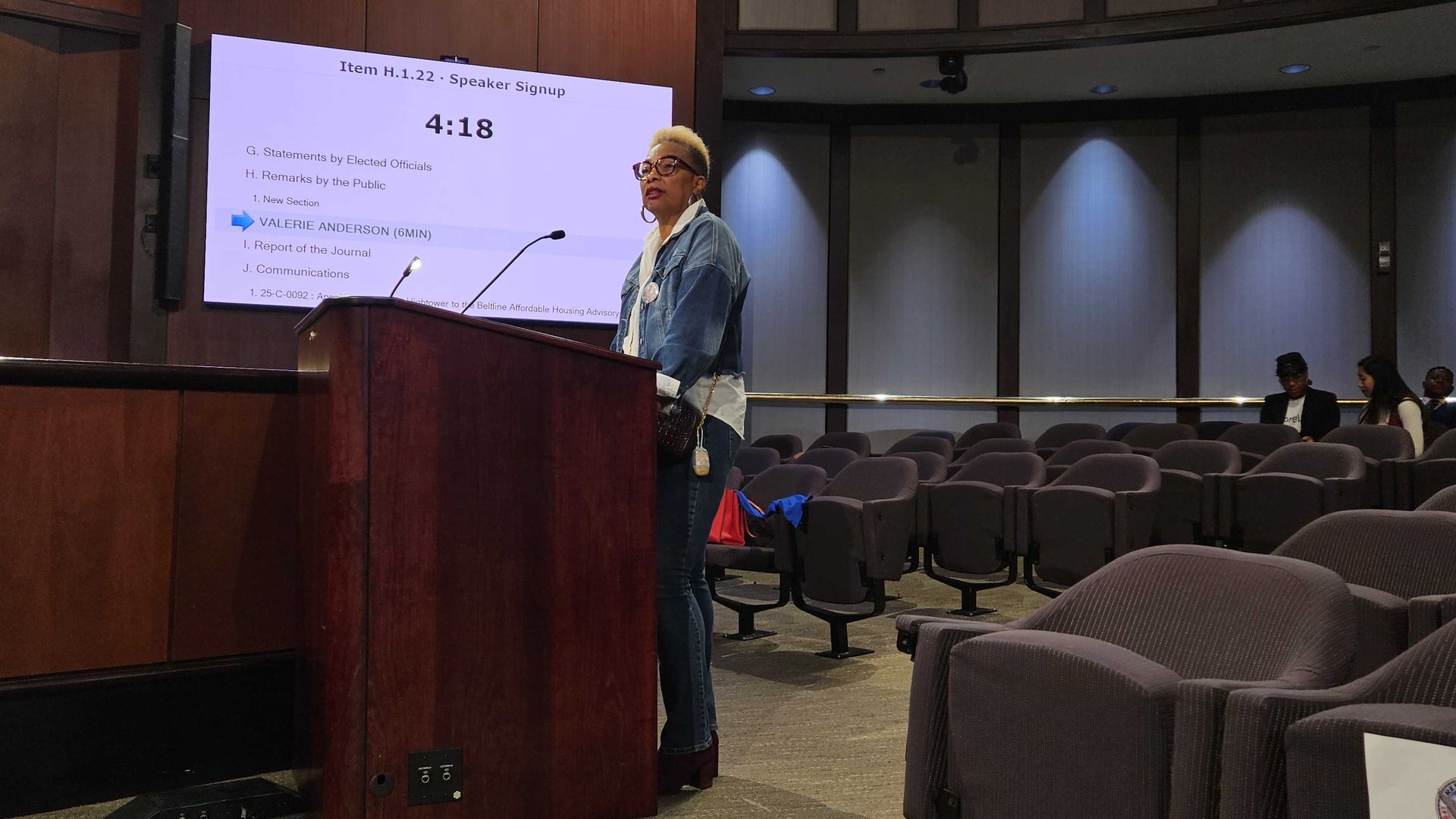 A woman in a denim jacket and glasses stands at a wooden podium in a meeting room with empty chairs. A screen behind shows a speaker signup and agenda with 4:18 time displayed.