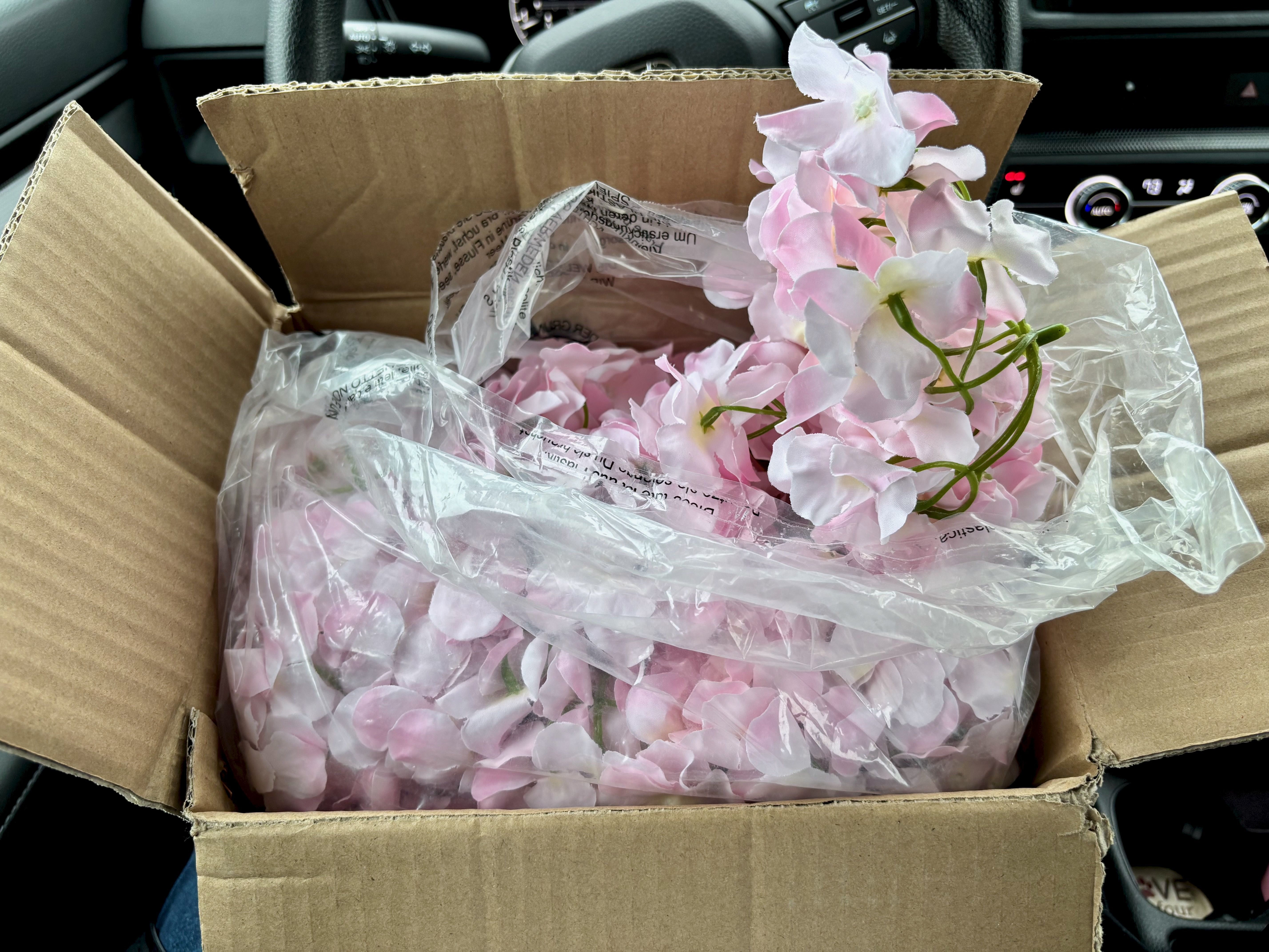 An open cardboard box inside a car containing light pink artificial flowers wrapped in clear plastic packaging.