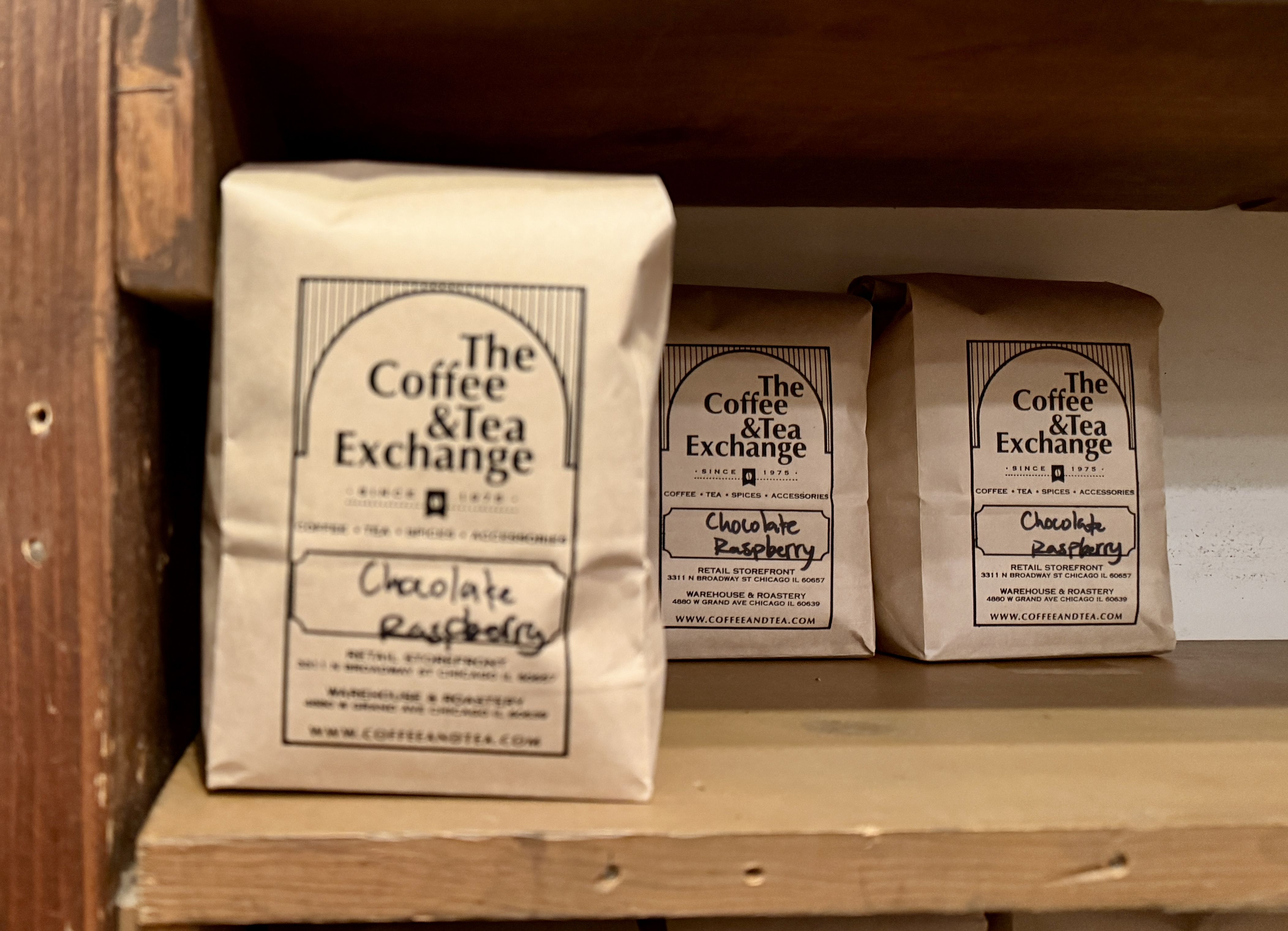 Three brown paper bags labeled The Coffee & Tea Exchange, all with handwritten chocolate raspberry flavor, placed on a wooden shelf.