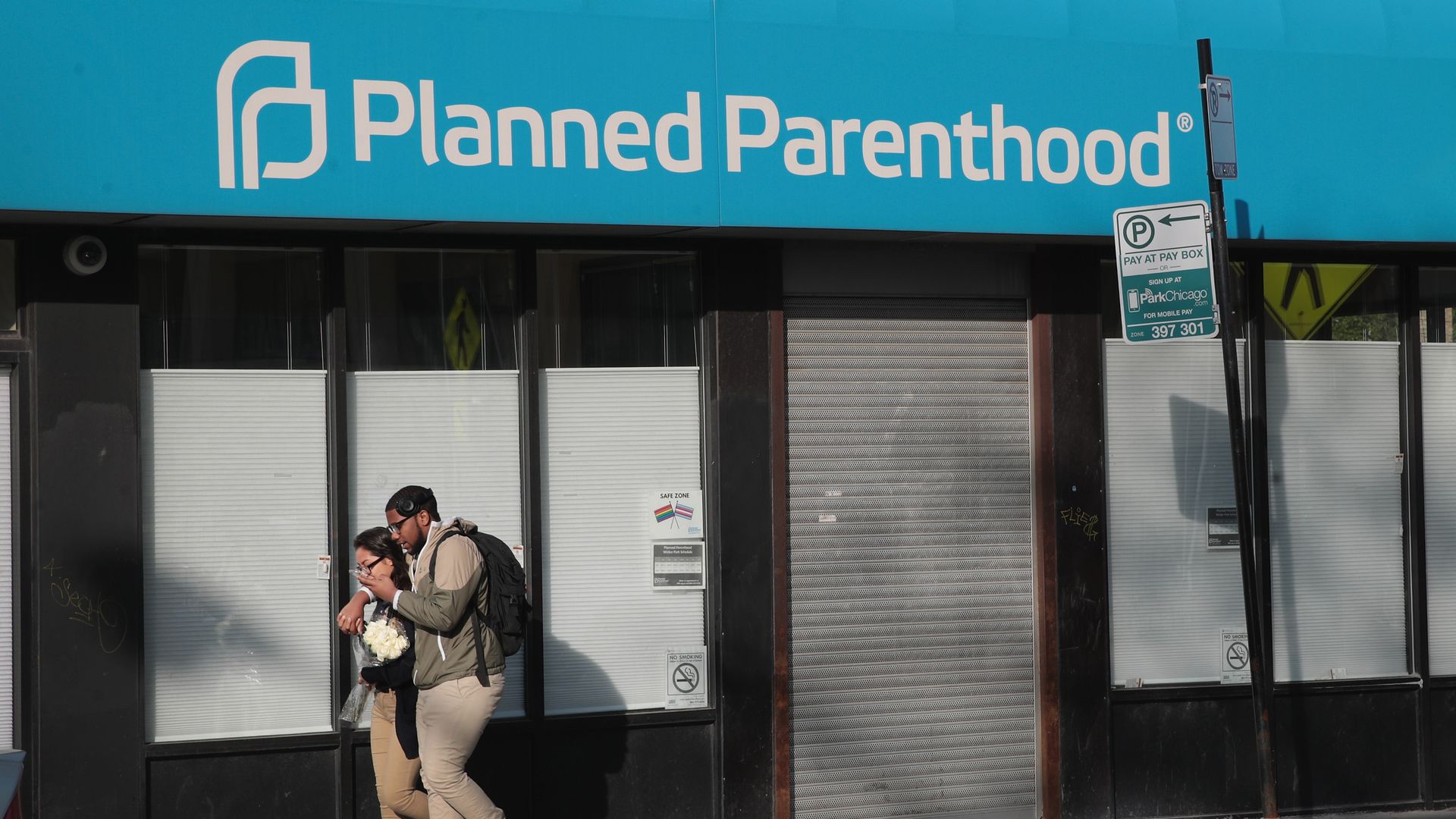 In this image, two people walk down the street in front of a blue awning with the white words "Planned Parenthood" printed on it.