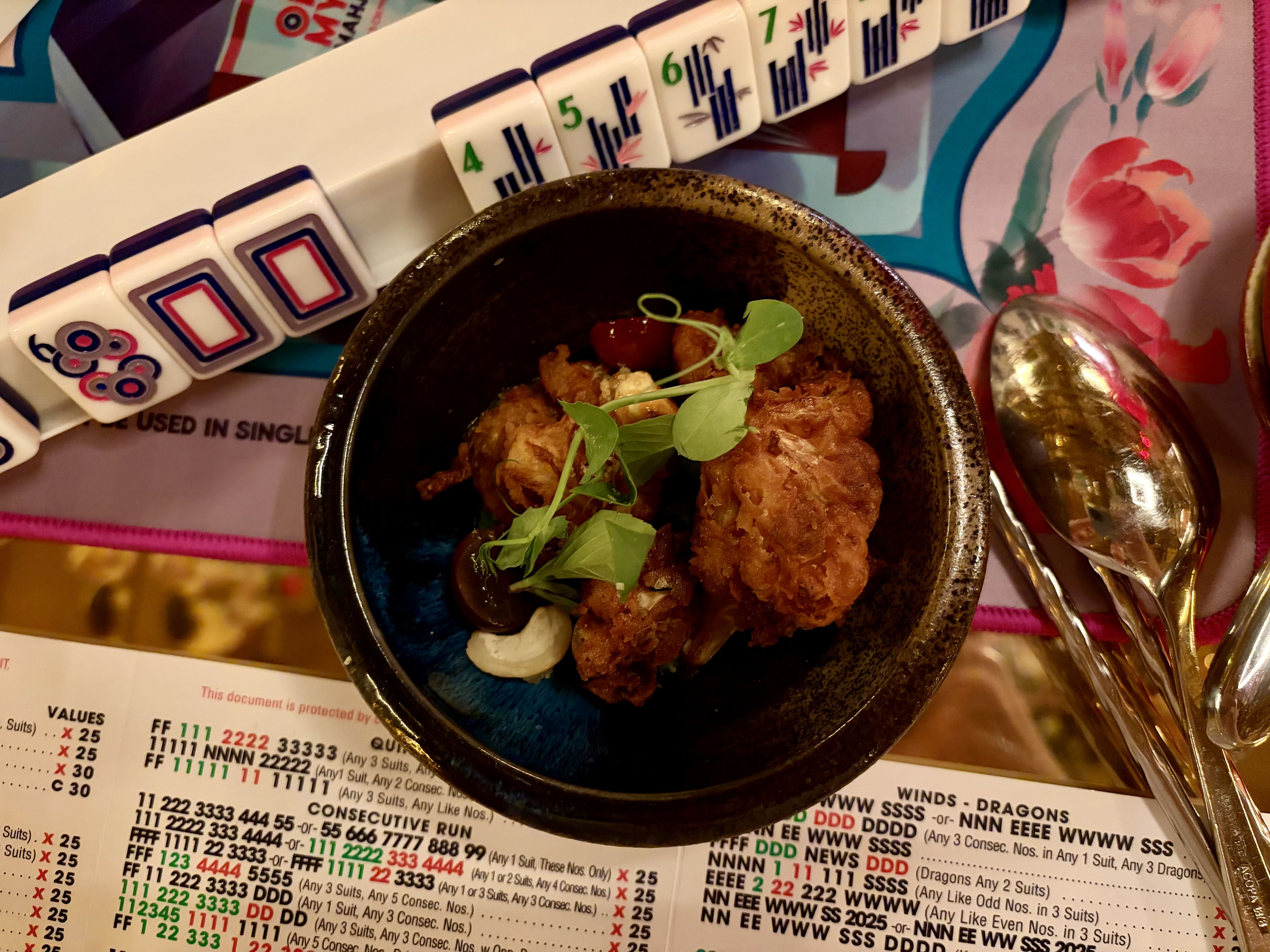 A dark ceramic bowl of fried cauliflower bites topped with greens sits on a colorful table beside a row of tiles, a shiny spoon, and a printed chart in the background.