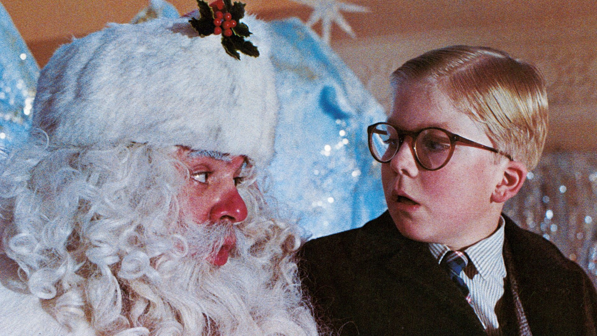 Scene from "A Christmas Story" showing Santa Claus with white curly hair, a red nose, and holly on hat, looking surprised at a young boy with round glasses and a brown coat.