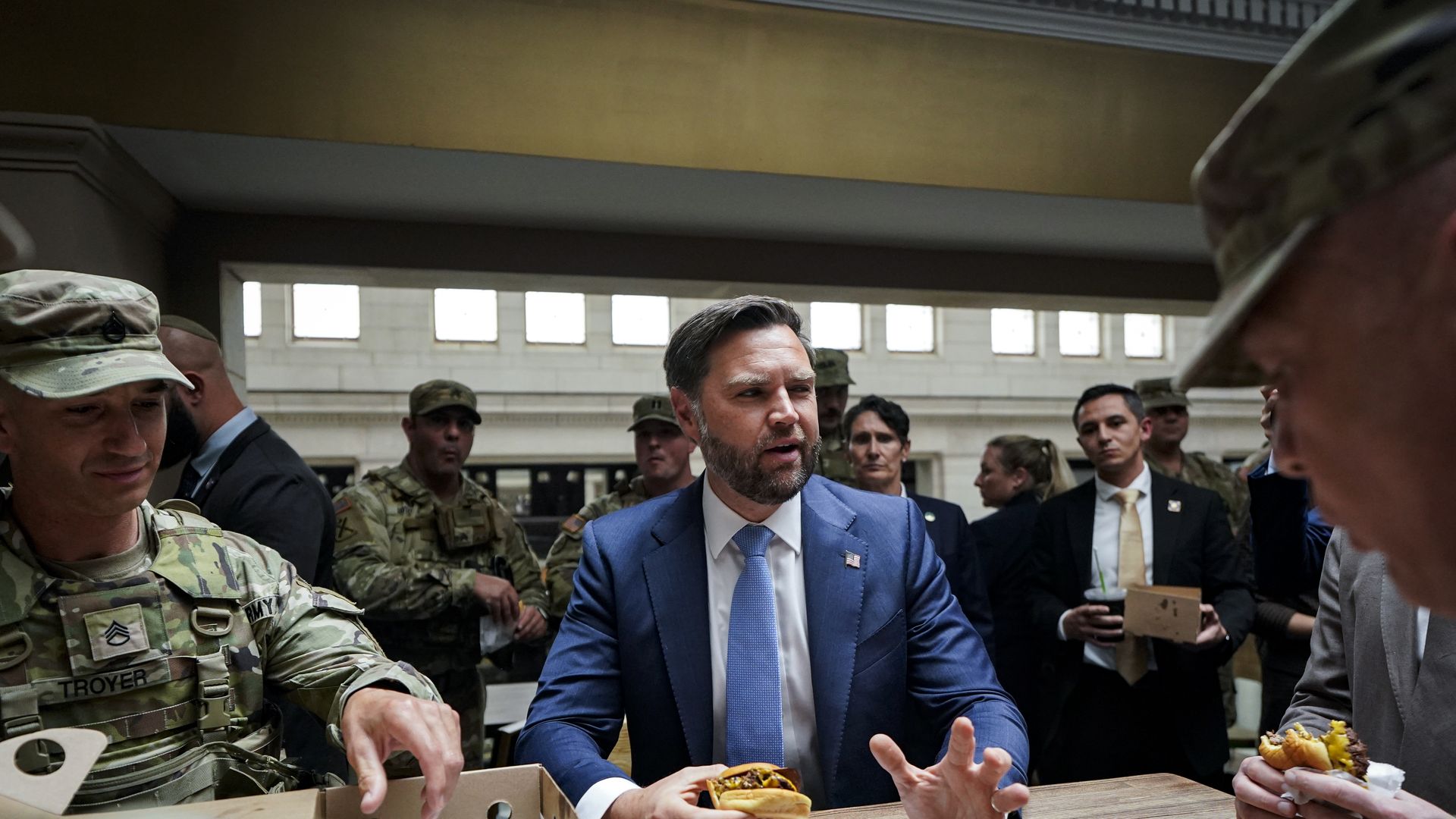 Man in blue suit and light blue tie holding a cheeseburger seated at a wooden table surrounded by U.S. Army soldiers in camouflage uniforms and others in formal attire indoors.
