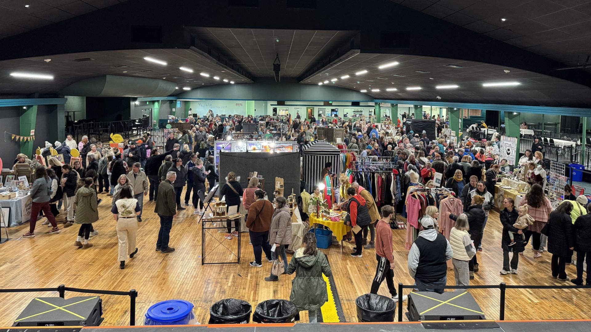Large indoor market with many people browsing booths selling clothes, art, and crafts under black ceiling with lights, wooden floor, and green walls.