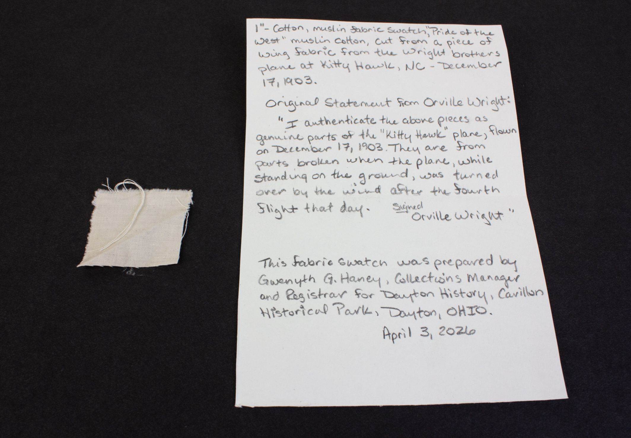 White handwritten note on a white sheet on a dark surface, with a small beige fabric swatch and string to the left; the note discusses a Wright brothers airplane and Kitty Hawk.