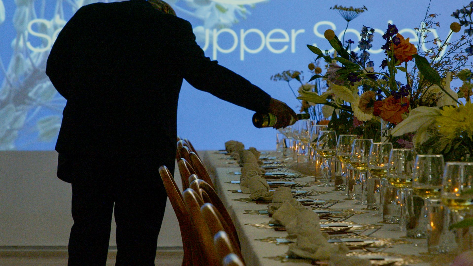 A pic of a person pouring wine into a glass that's set on a dinner table with multiple place settings