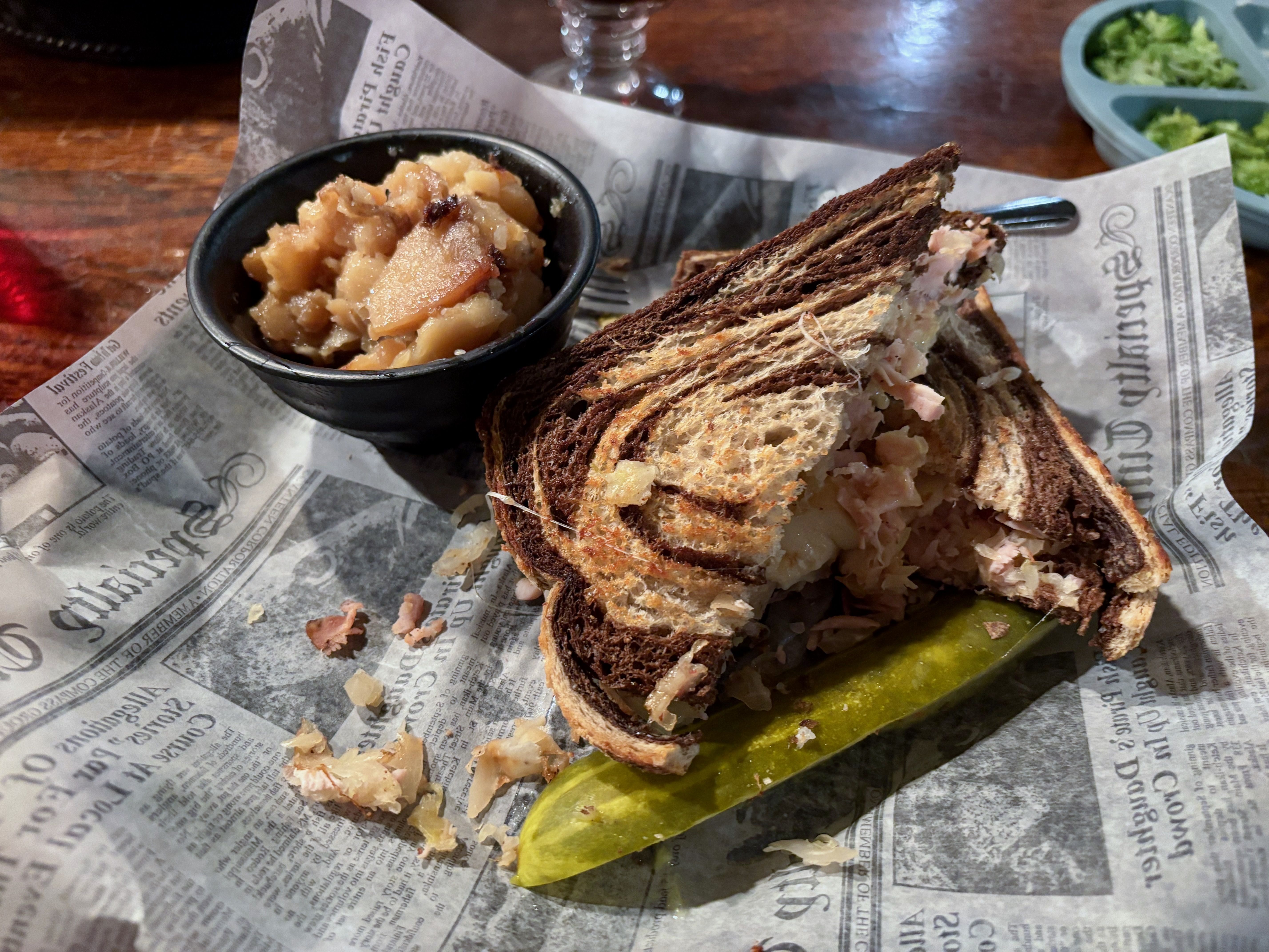 Rye marbled sandwich with sauerkraut and turkey, served on newspaper-style paper with a large dill pickle and a small black cup of chunky German potato salad.