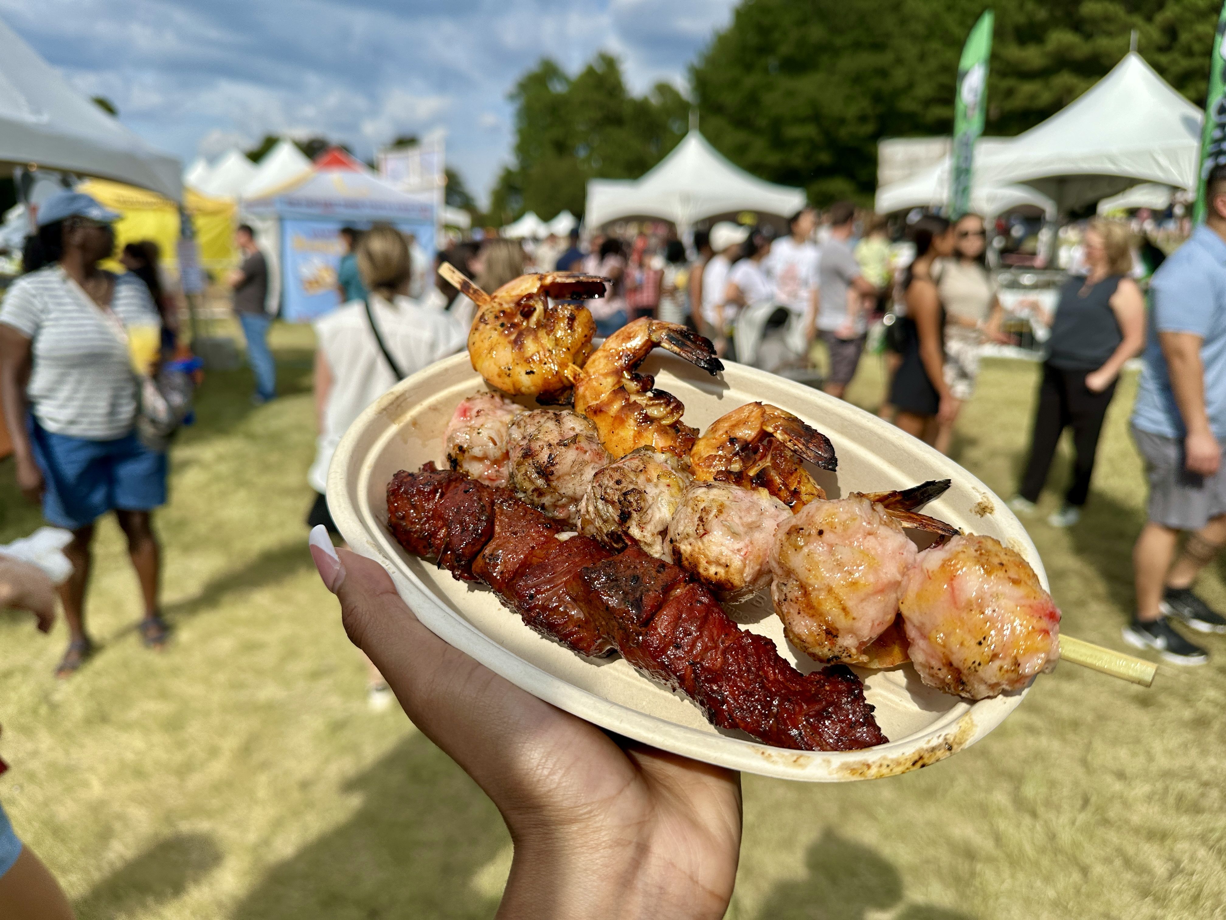 Hand holding a plate with skewers of grilled shrimp and meat at an outdoor food festival with tents and people in the background on a sunny day.