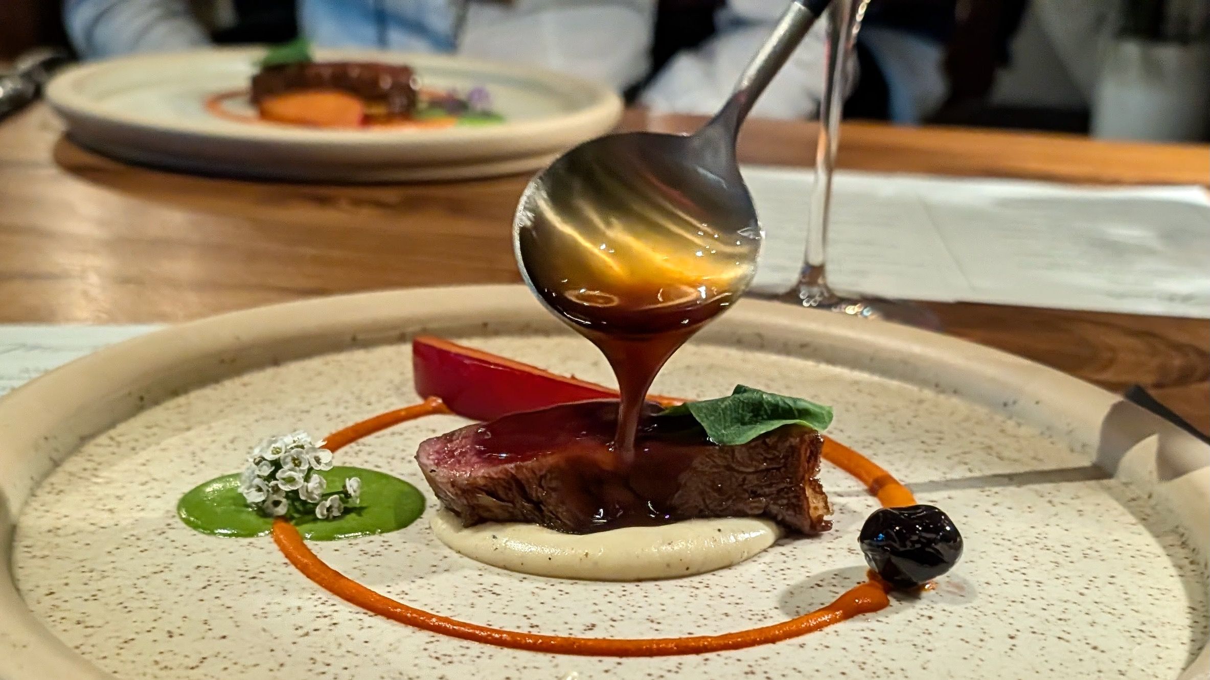 Close-up of a plated, fine-dining dish: a square of glazed steak with dark sauce flowing from a spoon, surrounded by green and orange sauces, a small leaf, and tiny white flowers on a speckled plate.