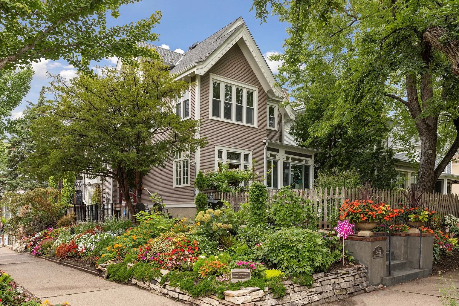 Two-story beige house with white trim behind a lush garden filled with colorful flowers and greenery, a wooden picket fence, and large leafy green trees under a partly cloudy blue sky.