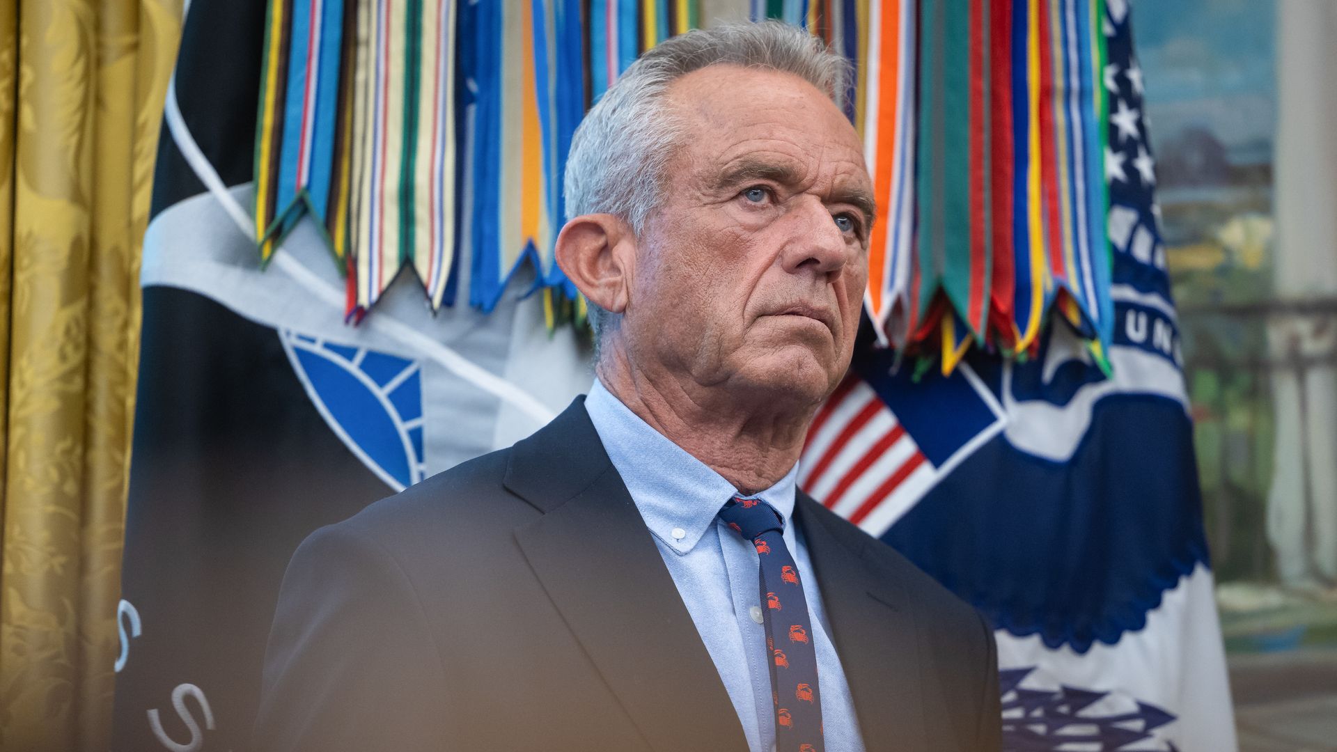 RFK Jr. looks on in the Oval Office while wearing a dark suit and a blue shirt.