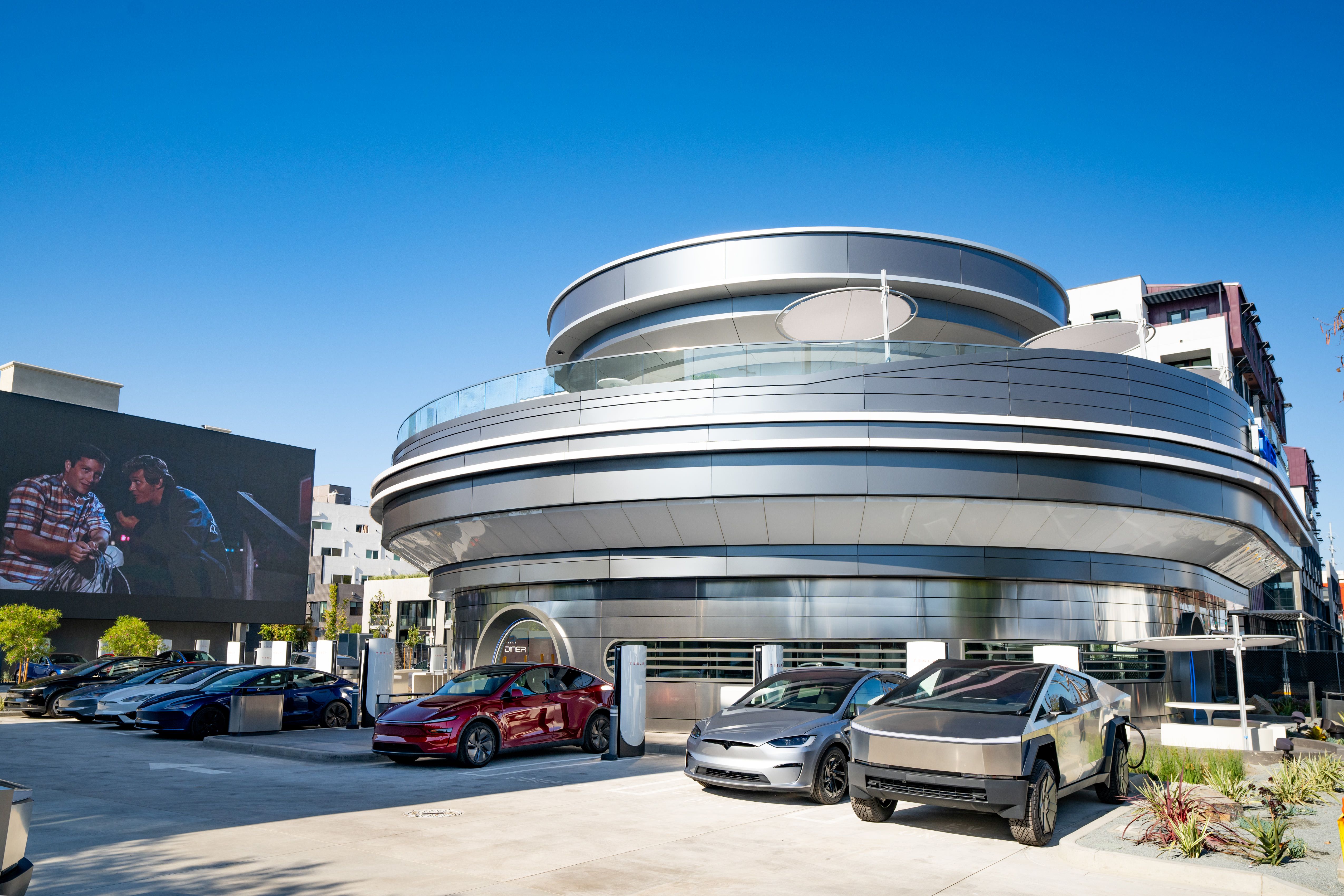 Tesla EVs parked and charging outside the newly opened Tesla Diner & Drive-In, a futuristic round building under a clear blue sky, with a large screen showing two people in conversation.