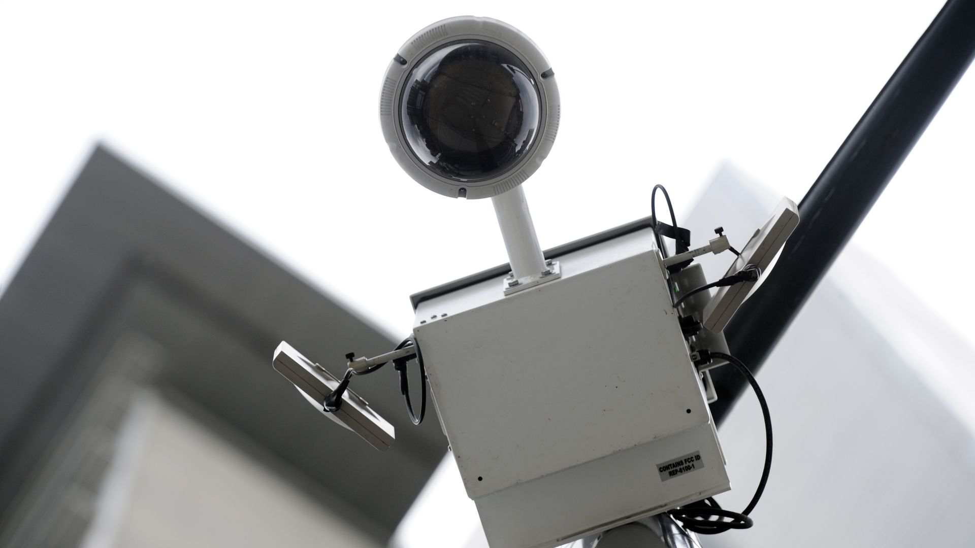 Close-up of a gray dome security camera on a metal pole with wiring, against a bright sky and blurred urban buildings in the background.