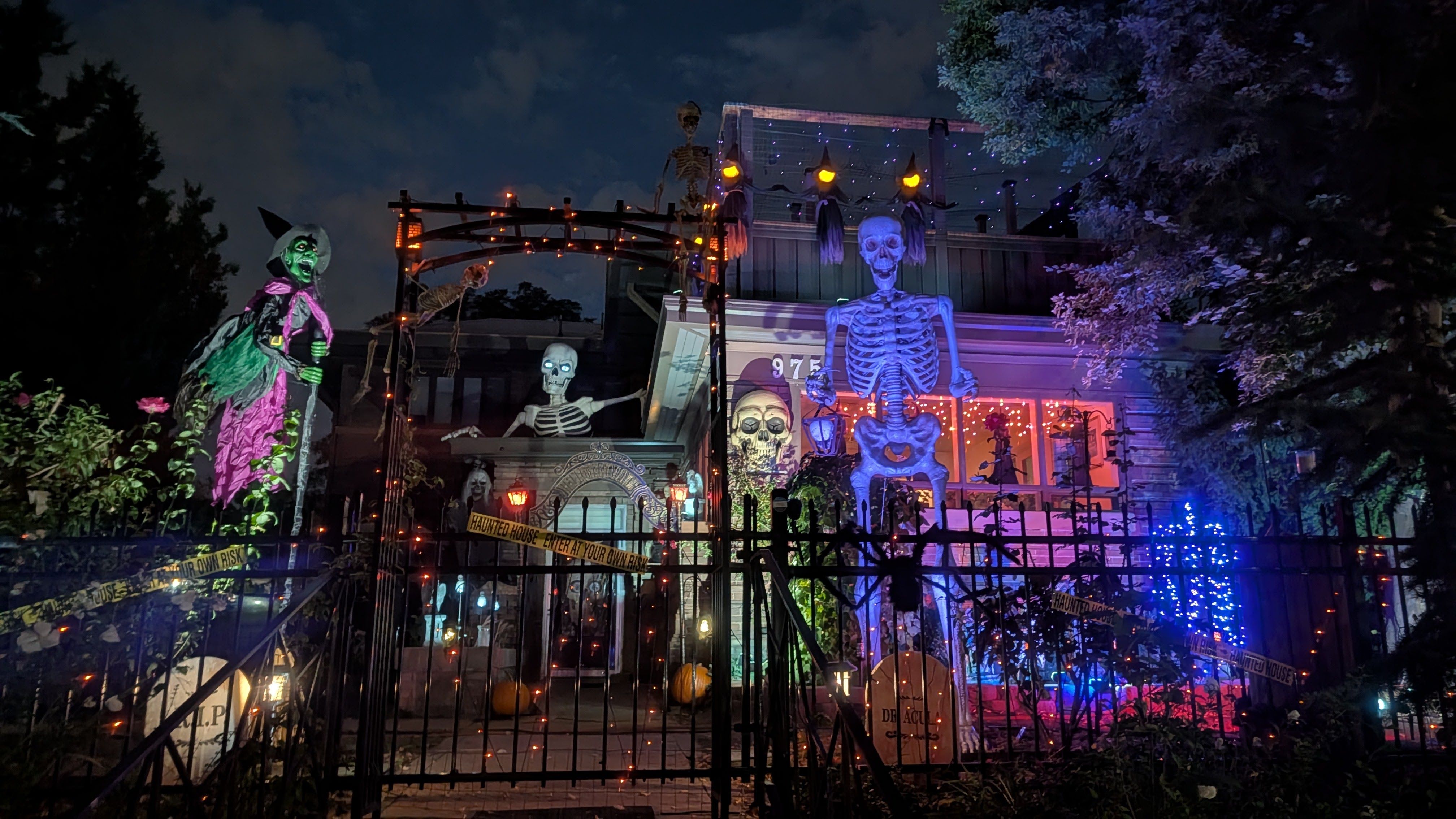 Nighttime haunted house scene with glowing skeletons, a witch figure, pumpkins, and string lights. A large blue skeleton sits on the balcony while a gate and yard glow purple and orange.