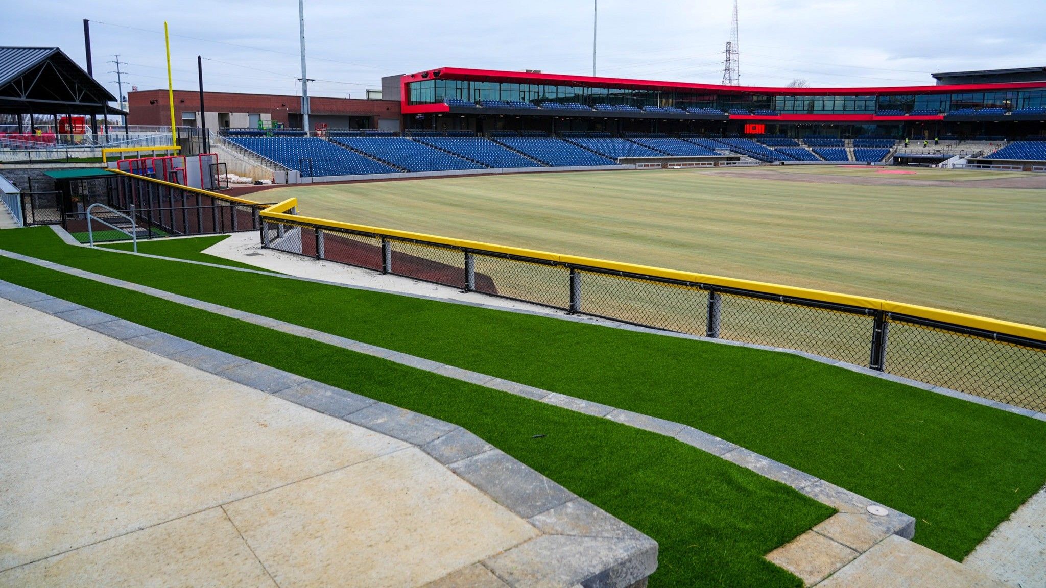Empty baseball field with green artificial turf, concrete seating area, blue stadium seats, and a red and black building under cloudy sky.