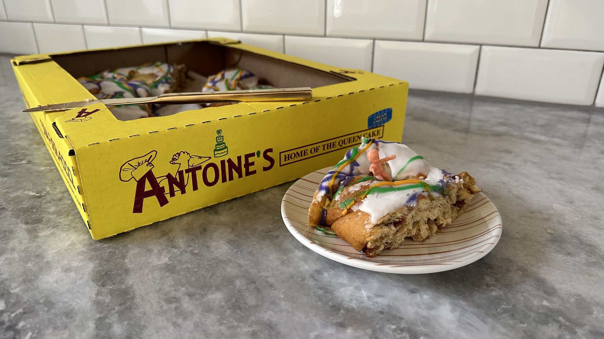 A slice of king cake sits on a small dessert plate with the baby tucked into the top. Behind the cake plate is a yellow box with the rest of the king cake. The box has a knife perched on top, and the box says Antoine's: Home of the Queen Cake.