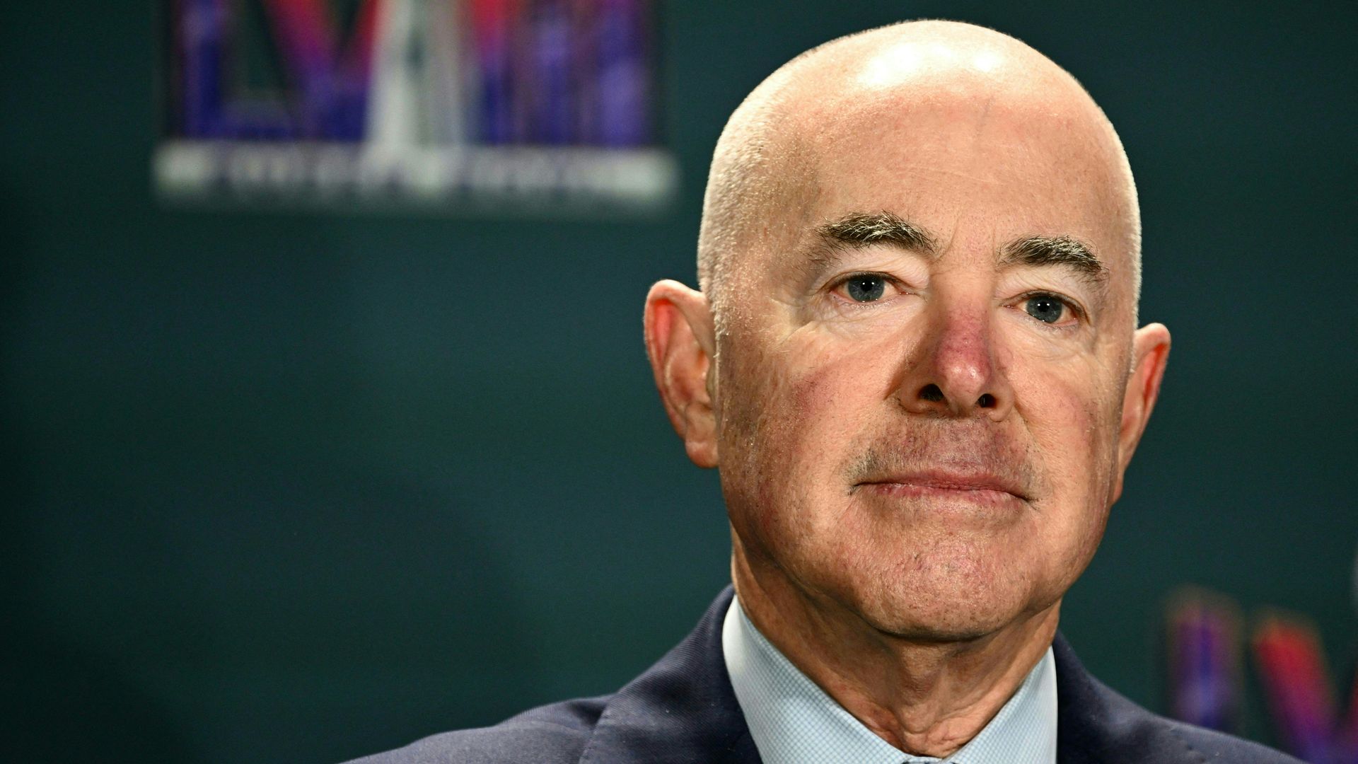 Alejandro Mayorkas, wearing a blue suit and light teal shirt against a turquoise Superbowl LVIII backdrop 