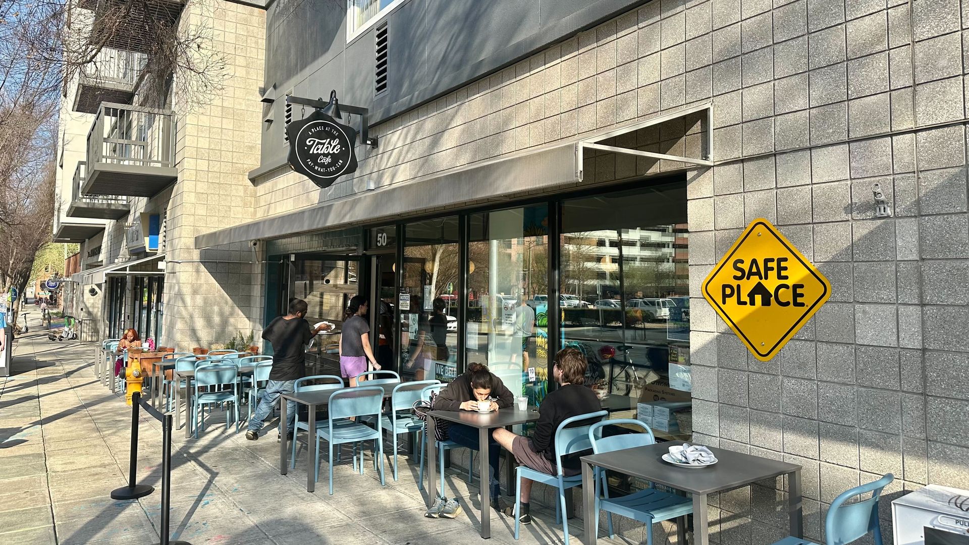 Outdoor cafe on a sunny sidewalk with light-blue chairs and gray tables along a gray-tiled building. A round Table Cafe sign hangs above; a yellow "SAFE PLACE" sign is on the wall as customers sit and eat.
