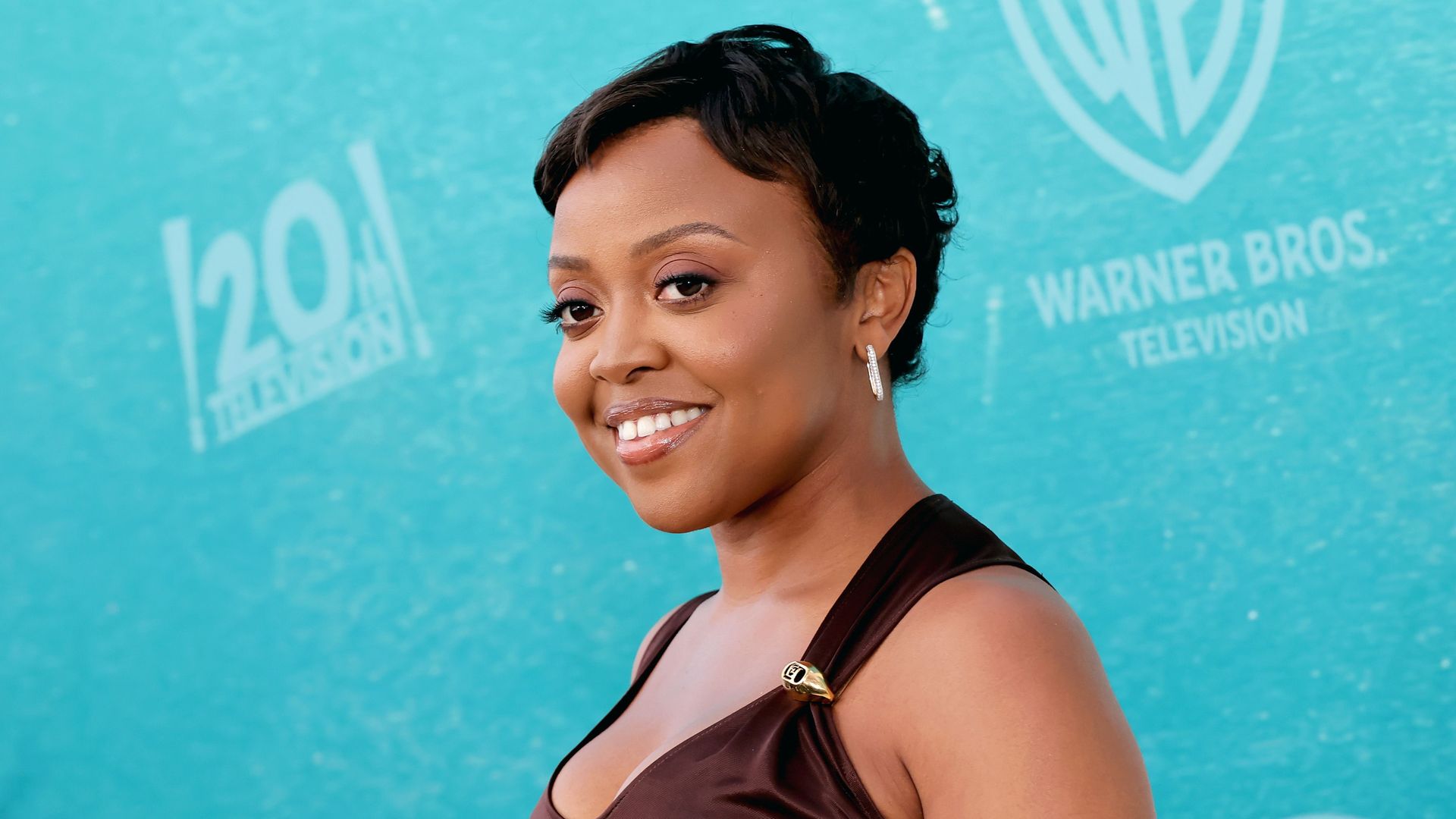 Portrait of a smiling Black woman with short black hair, wearing a brown dress and silver earrings, posed against a turquoise backdrop with a Warner Bros. Television logo.