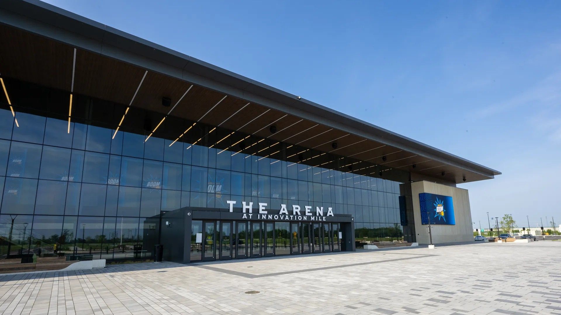 Modern glass-front arena with a long overhanging roof and large glass doors. The facade reads "THE ARENA AT INNOVATION MILE" in white letters; blue sky and a paved plaza surround it.