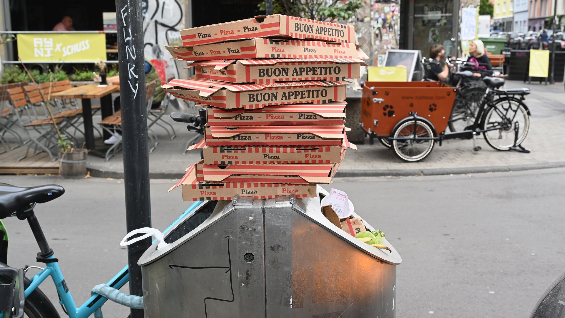Stack of pizza boxes labeled BUON APPETITO sits on a gray street vendor counter in a busy urban scene, with bicycles, outdoor tables, graffiti-covered pole, and colorful buildings behind.