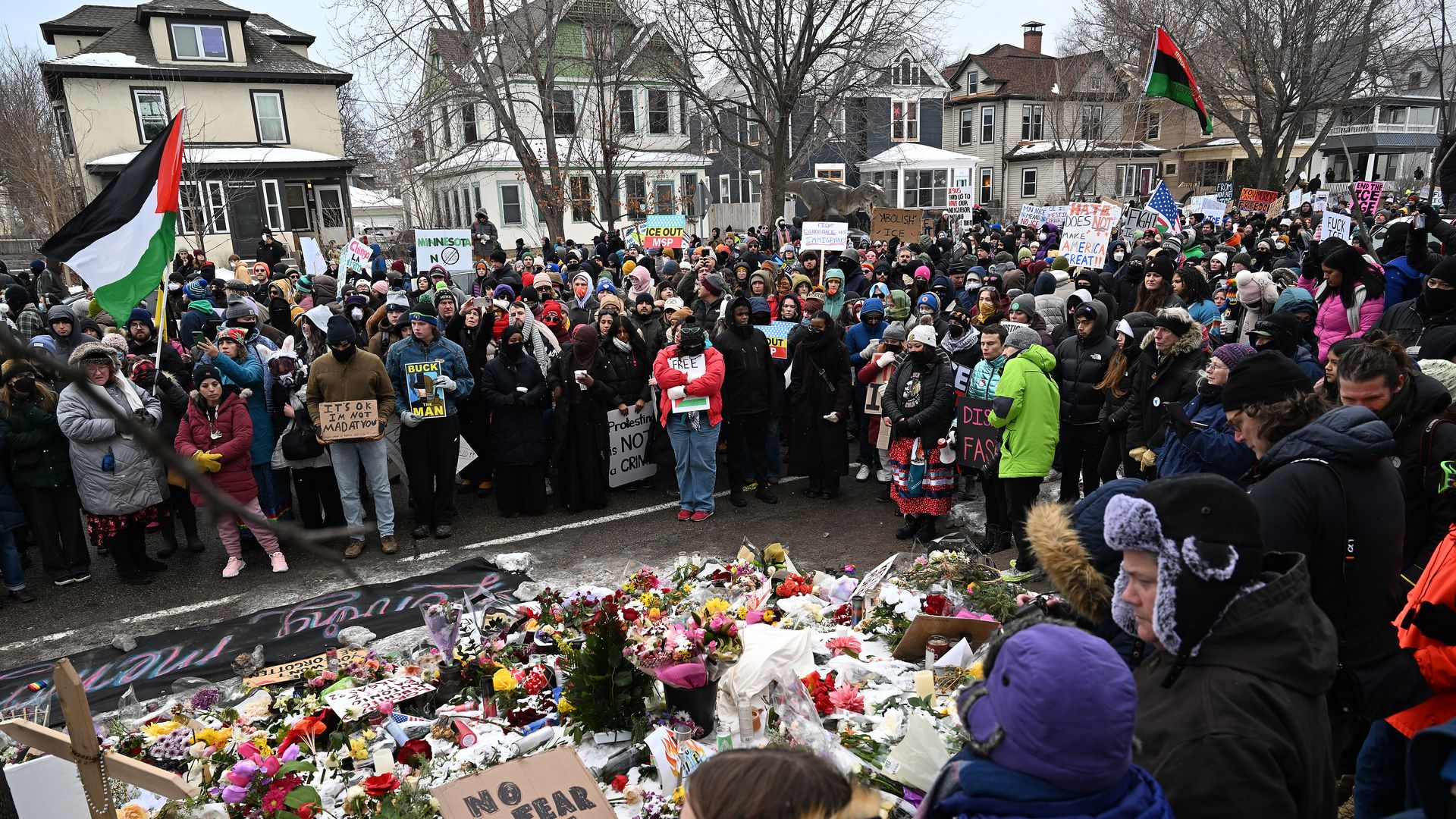 Large crowd gathered outdoors in winter near houses, holding signs and flags, surrounding a memorial with flowers and candles, including a sign saying "NO FEAR LOVE WINS".