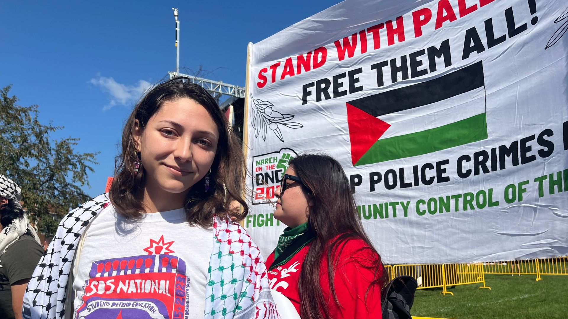 A woman wearing a T-shirt that says "SDS National" wearing a geen, red and black Palestinian keffiyeh next to a sign that reads "STAND WITH PALESTINE FREE THEM ALL!" with a Palestinian flag.