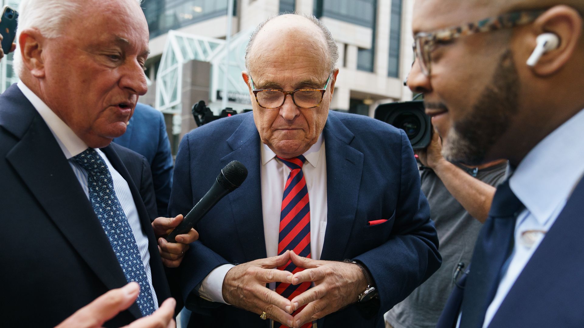Rudy Giuiliani stands between two men and looks down with his fingers touching 