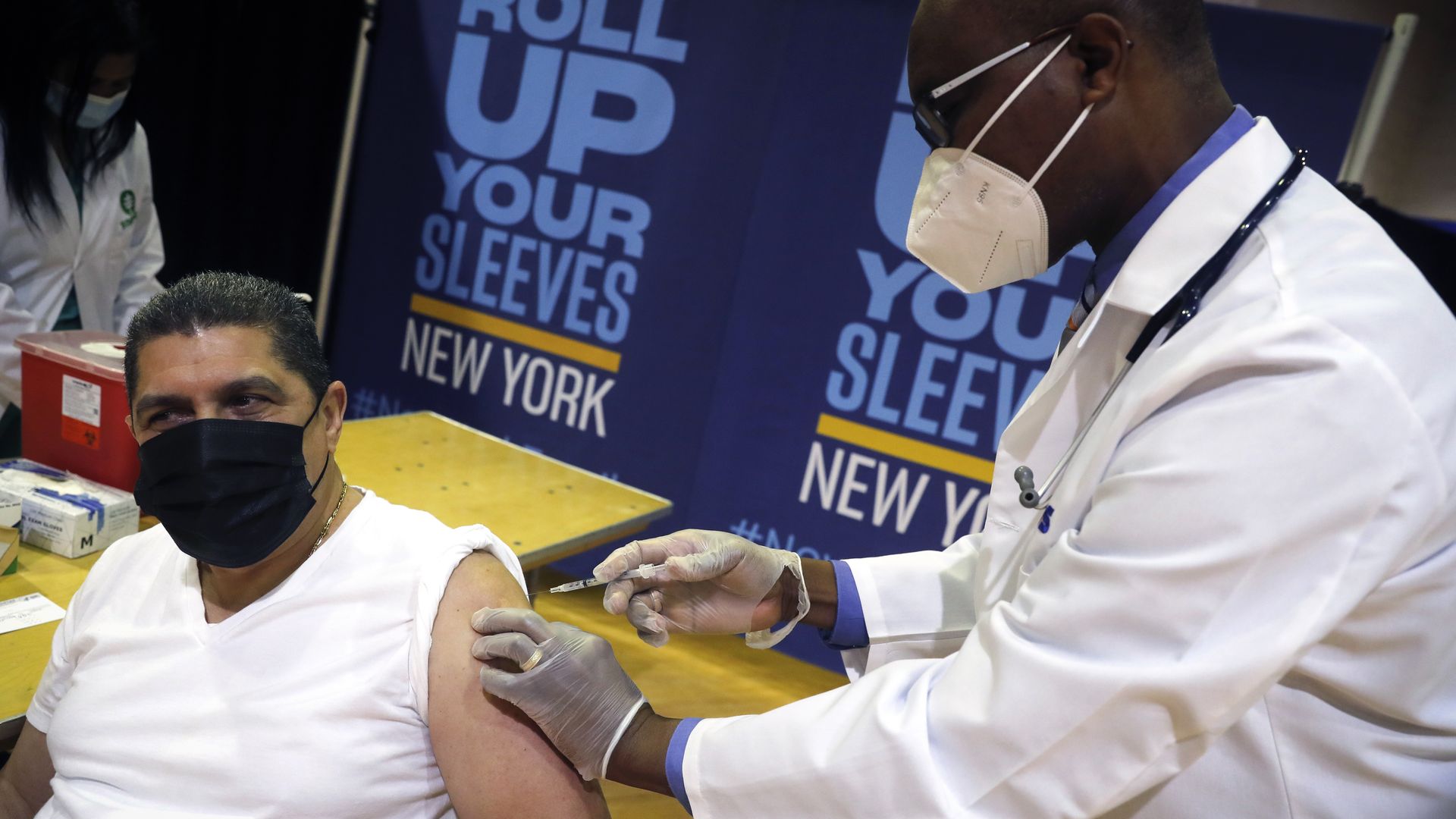 A doctor gives a vaccination to a man who is sitting in a chair with his sleeve rolled up.