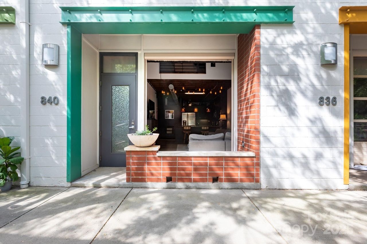 Front of a modern building with a dark gray door and large open window framed by red bricks and green trim, showing a living room with gray couch and hanging lights inside.
