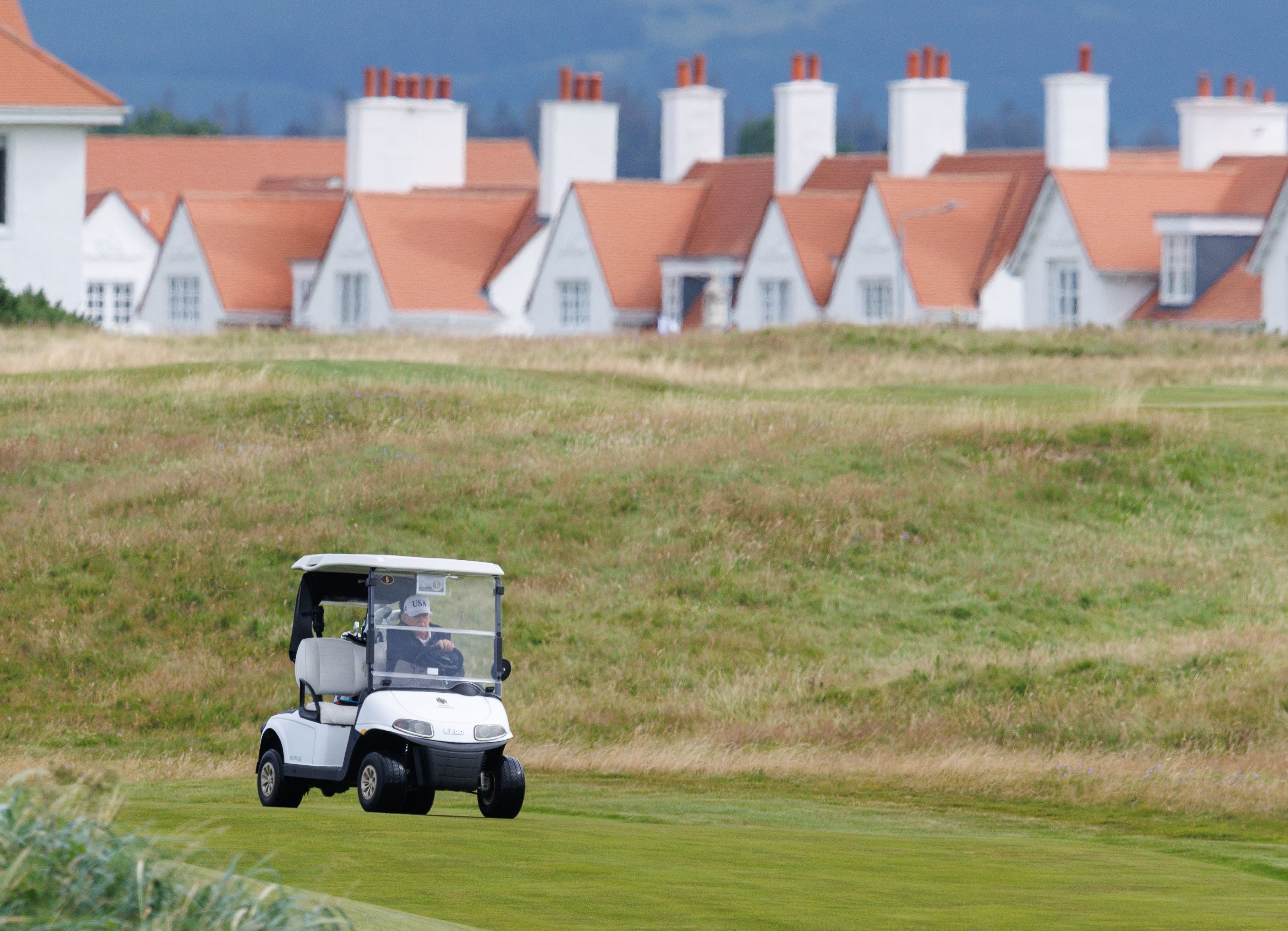 Person driving a white golf cart on a green golf course with tall grass and a row of white houses with orange roofs and chimneys in the background under a cloudy sky.