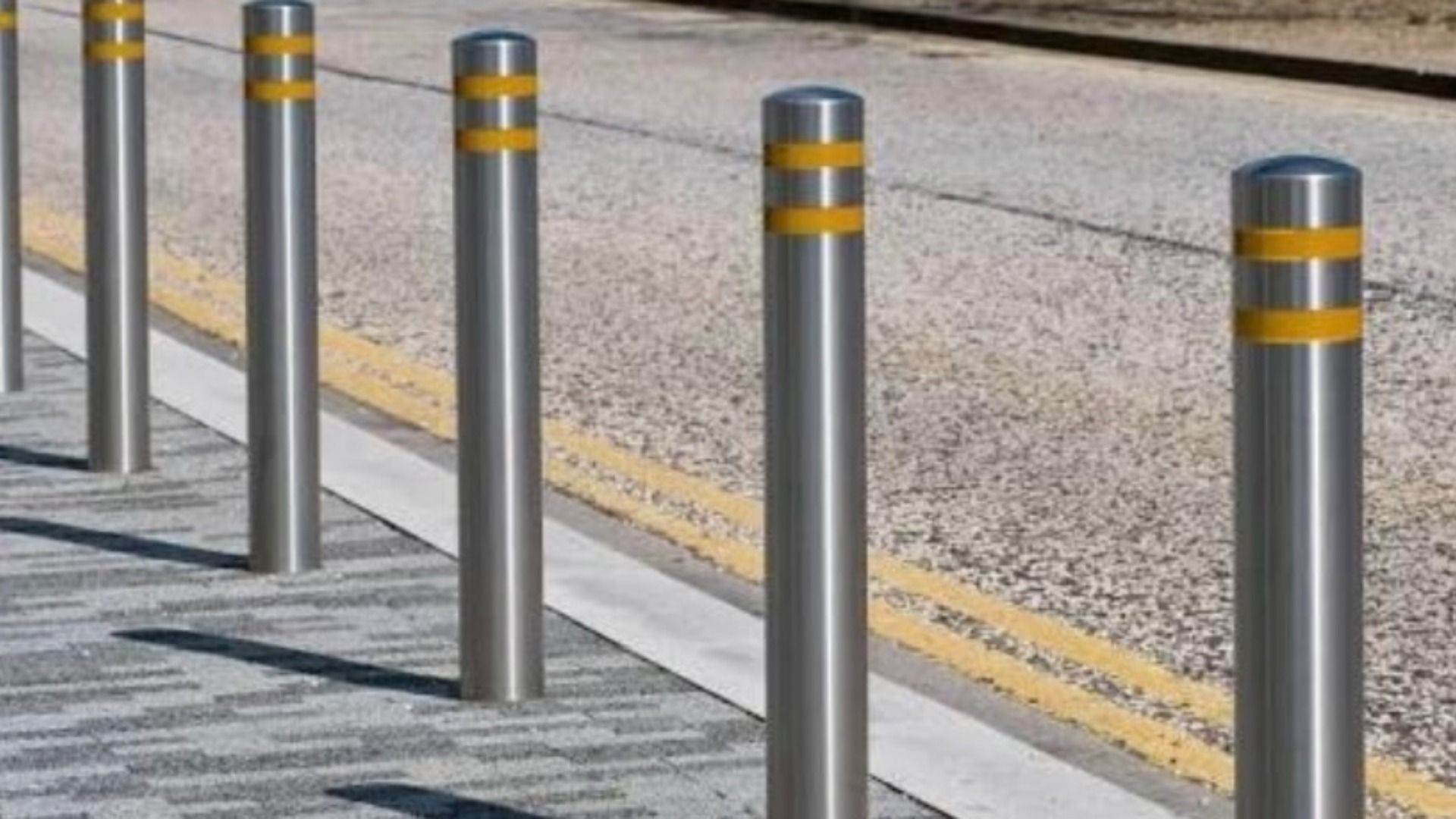 Row of stainless steel bollards with yellow bands line a sidewalk, forming a protective barrier beside a gravel road.