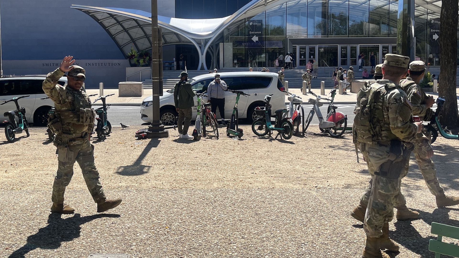 Soldiers in camo uniforms walking and waving near a bike and scooter rack outside the Smithsonian Institution on a sunny day.