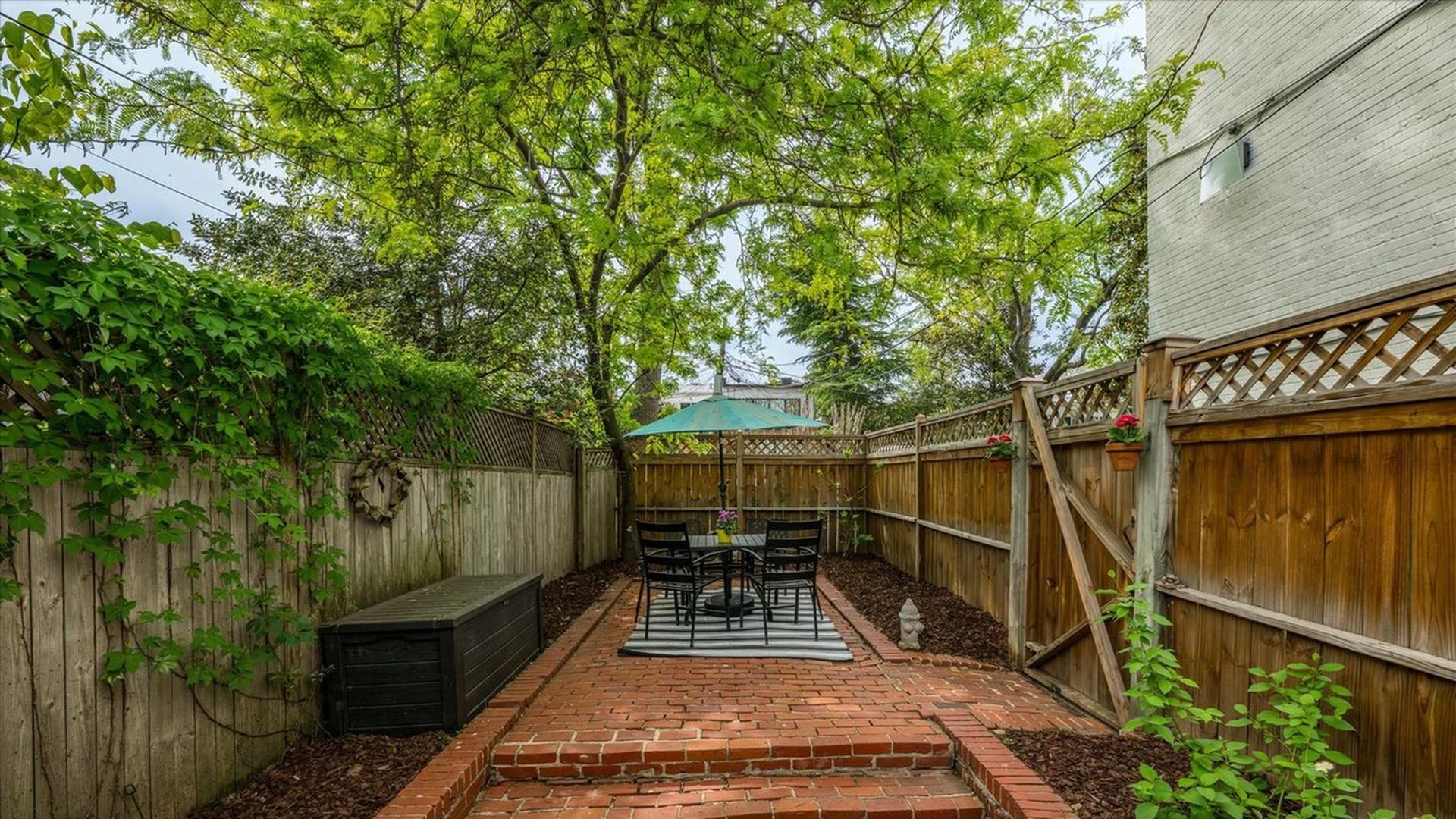 Backyard patio with red brick steps and a central dining set under a green umbrella, surrounded by wooden fences and leafy vines; a black storage box sits to the left.