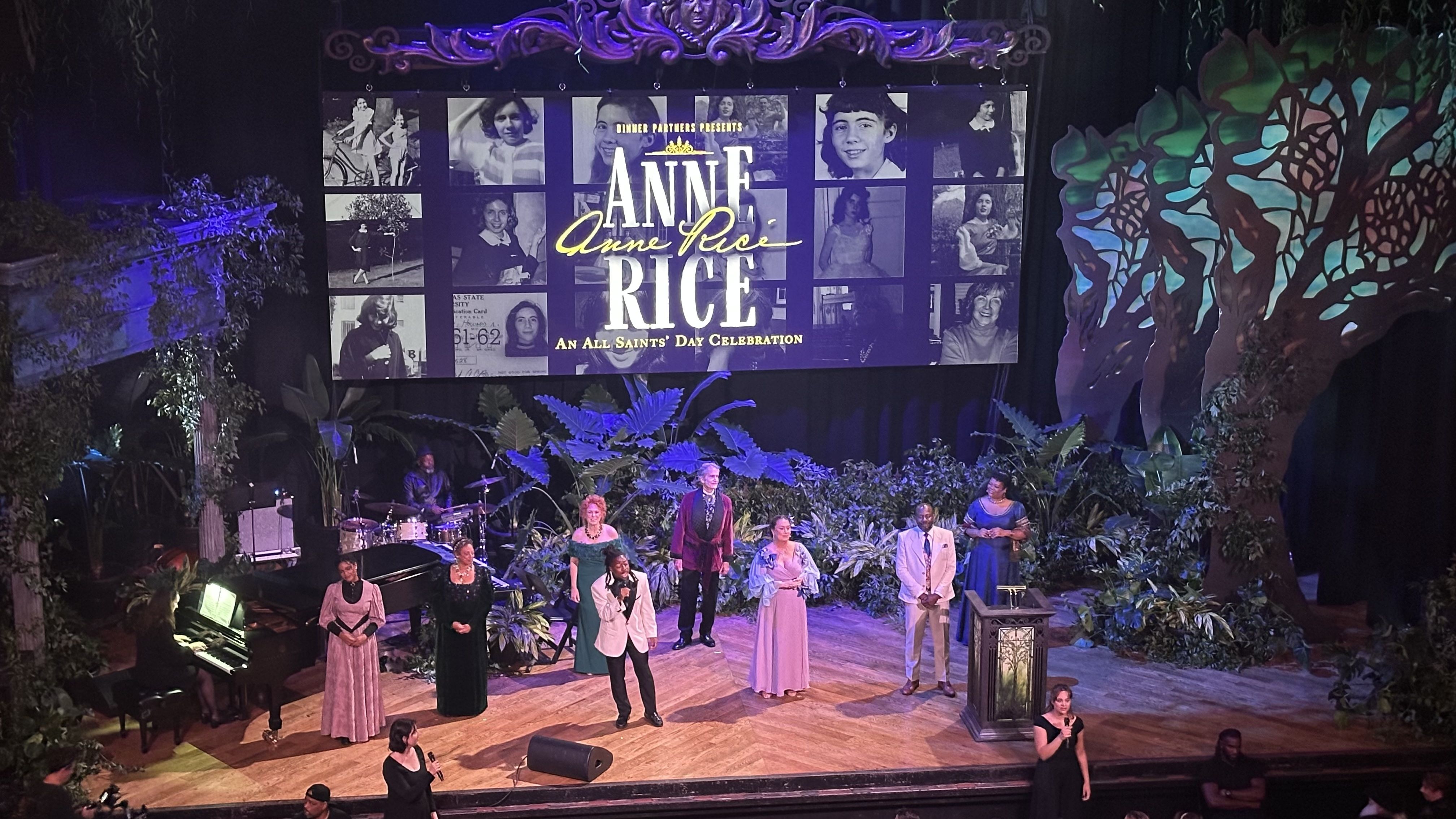 Stage scene with performers in vintage attire and a pianist, surrounded by green plants and large decorative trees, with a screen displaying "Anne Rice: An All Saints' Day Celebration".