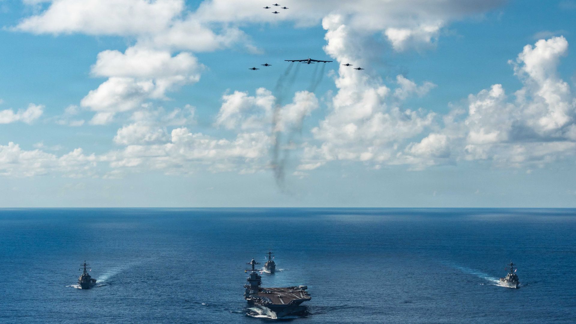 The U.S. Navy's Gerald R. Ford Carrier Strike Group, including the flagship USS Gerald R. Ford, front, operates as a joint, multi-domain force with a U.S. Air Force B-52 Stratofortress, over a blue sea and in a blue sky with fluffy, white clouds on Nov. 13.