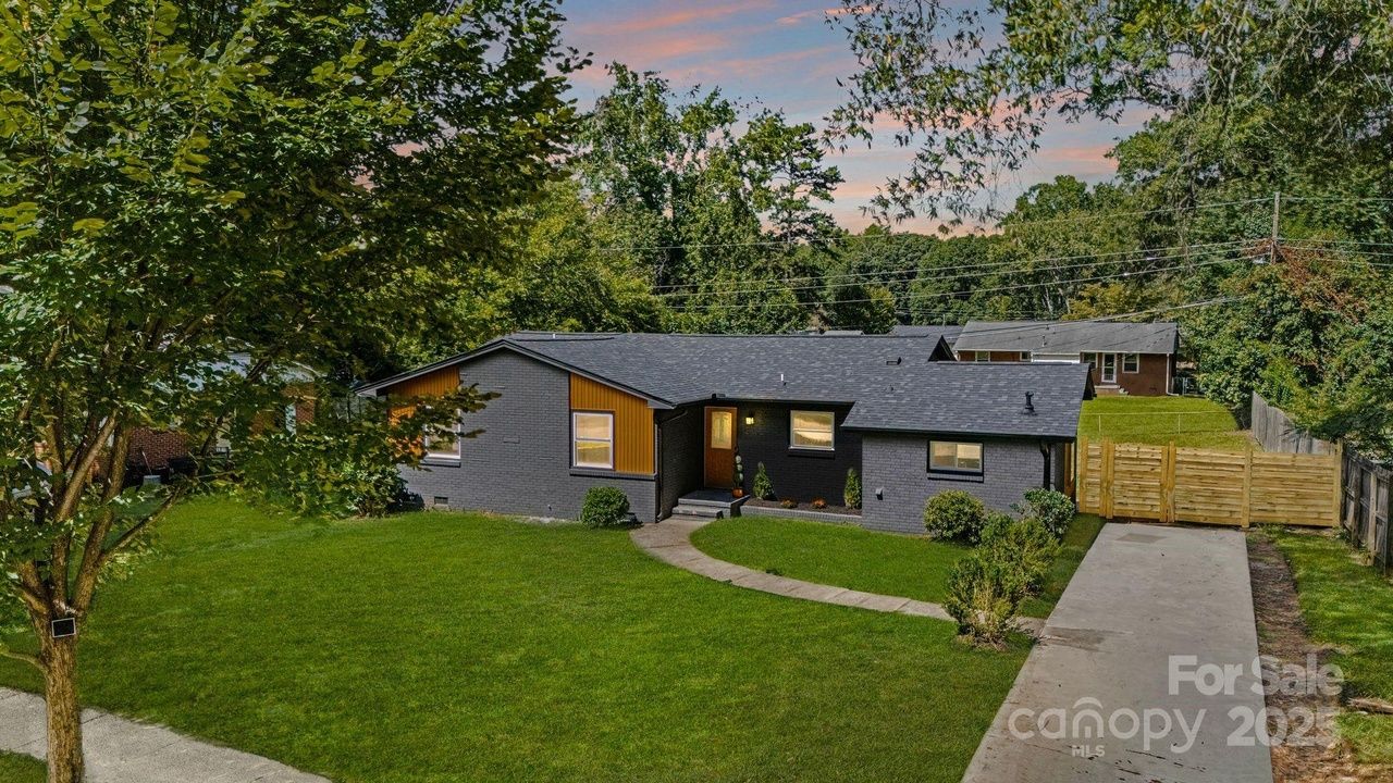 Single-story gray brick house with mustard yellow accent, black roof, front porch light on, concrete driveway, green lawn, trees, and wooden fence under a pink and blue sky at sunset.