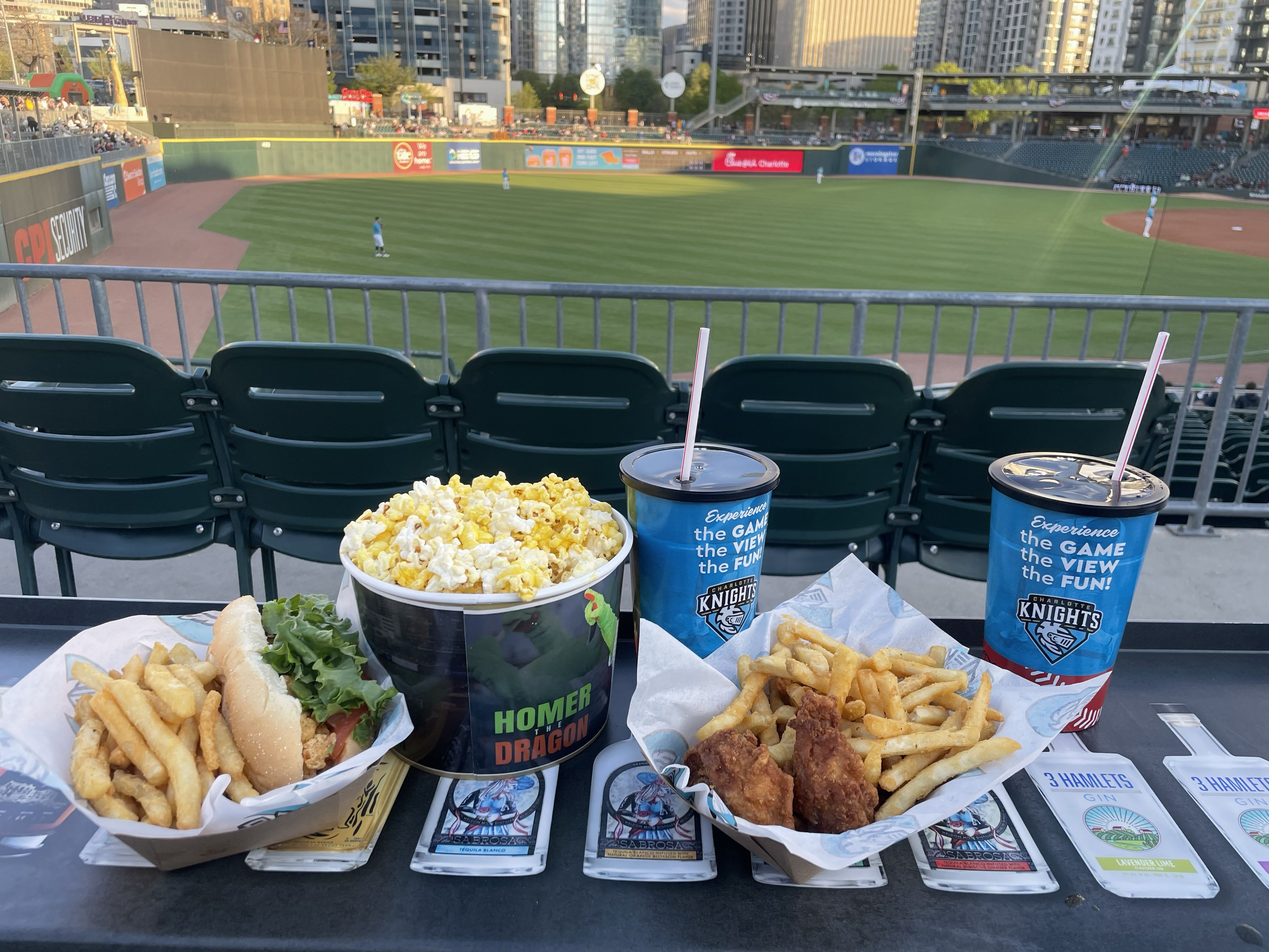 Food and drinks including: a popcorn bucket, a sandwich with fries, fried chicken with fries, and two fountain sodas; in front of a baseball stadium field.