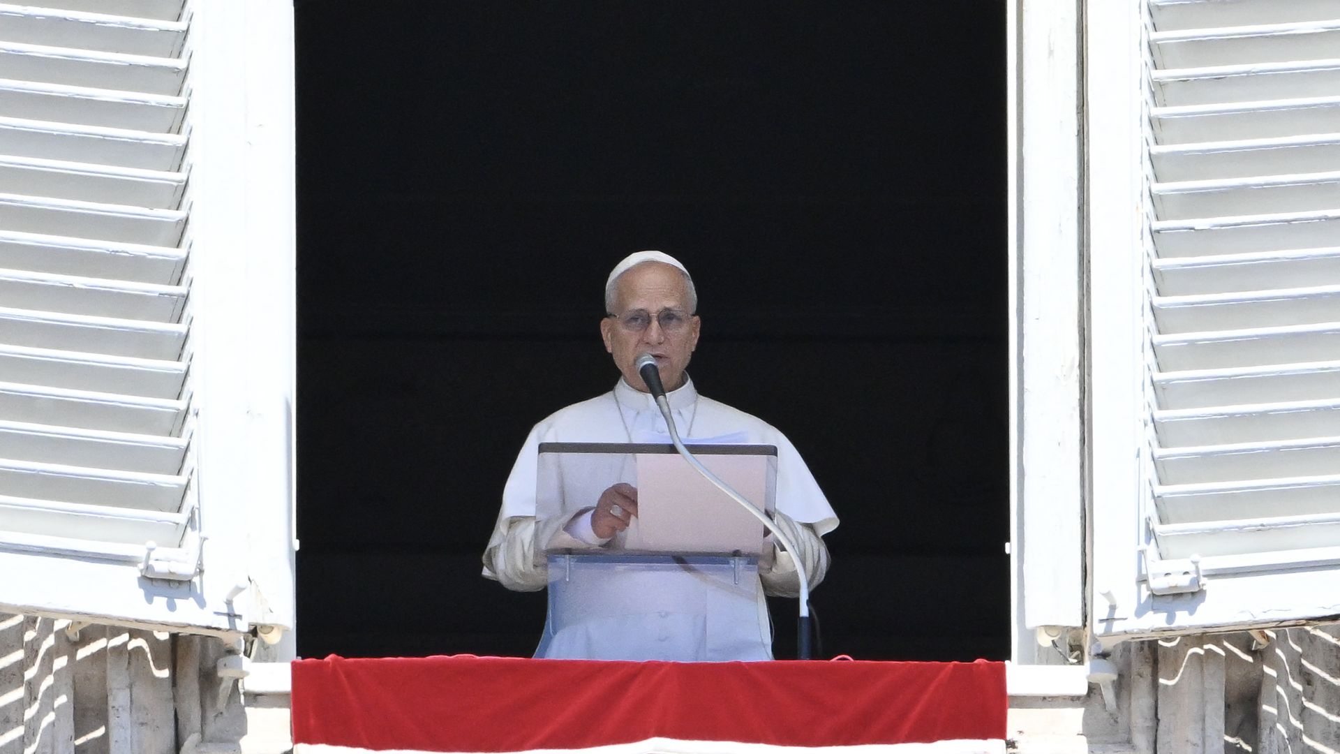 Pope Leo XIV, wearing a white cap and white cassock, speaks into a microphone at an open window with white shutters and draped in a red flag.
