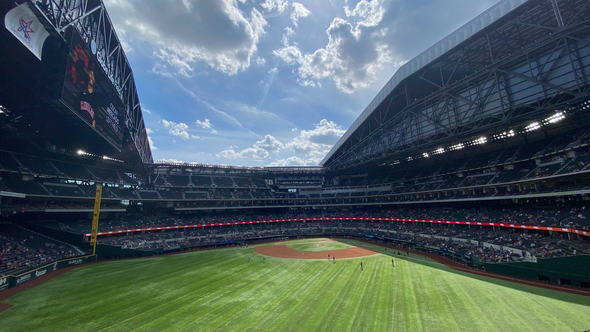 Globe Life park on a sunny day. The sun peeks out from behind the clouds, over the green outfield.