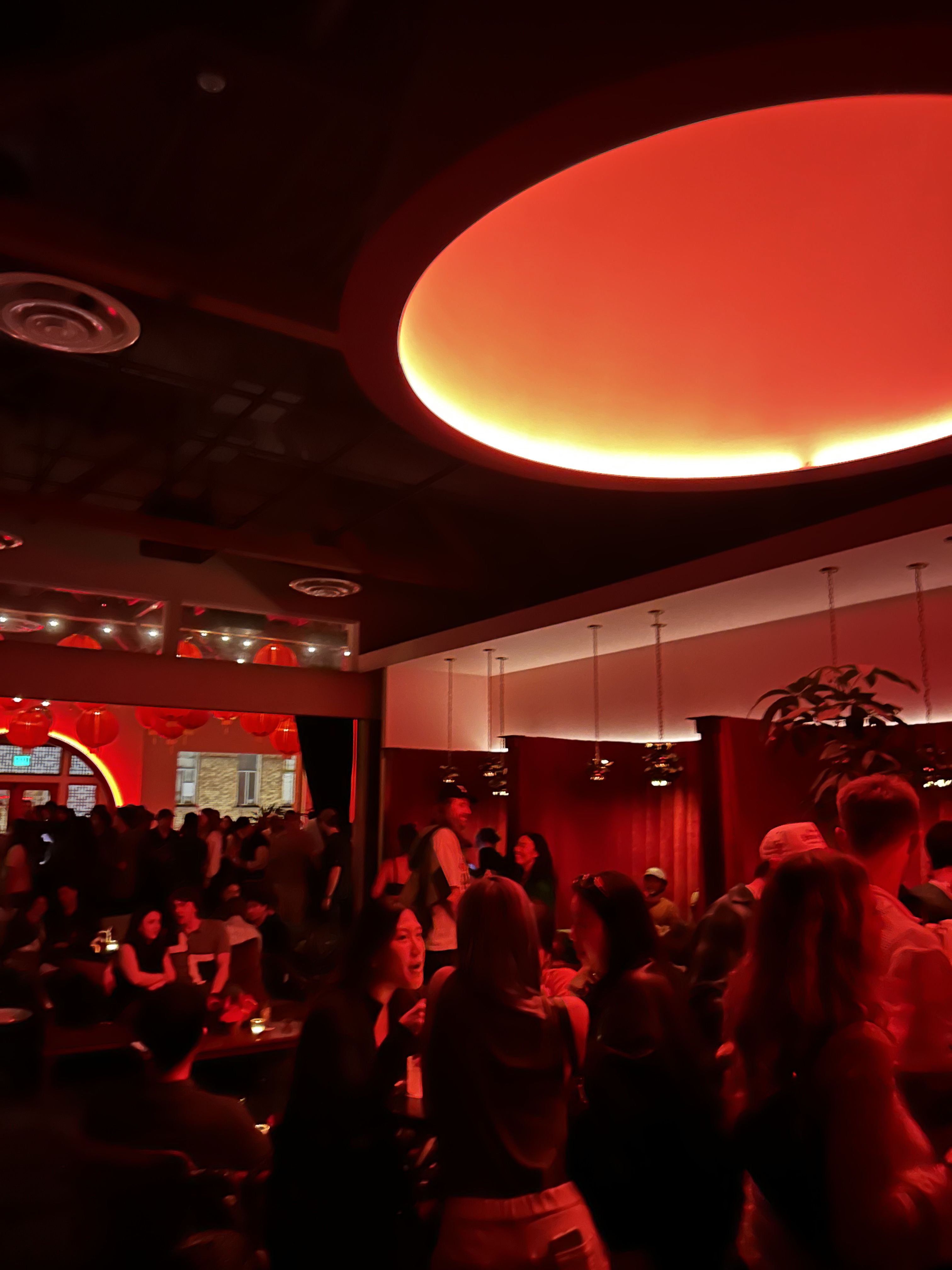 Red-lit nightclub or bar interior with a large circular orange ceiling light. A crowded room of people socializing, standing and sitting at tables along the walls.