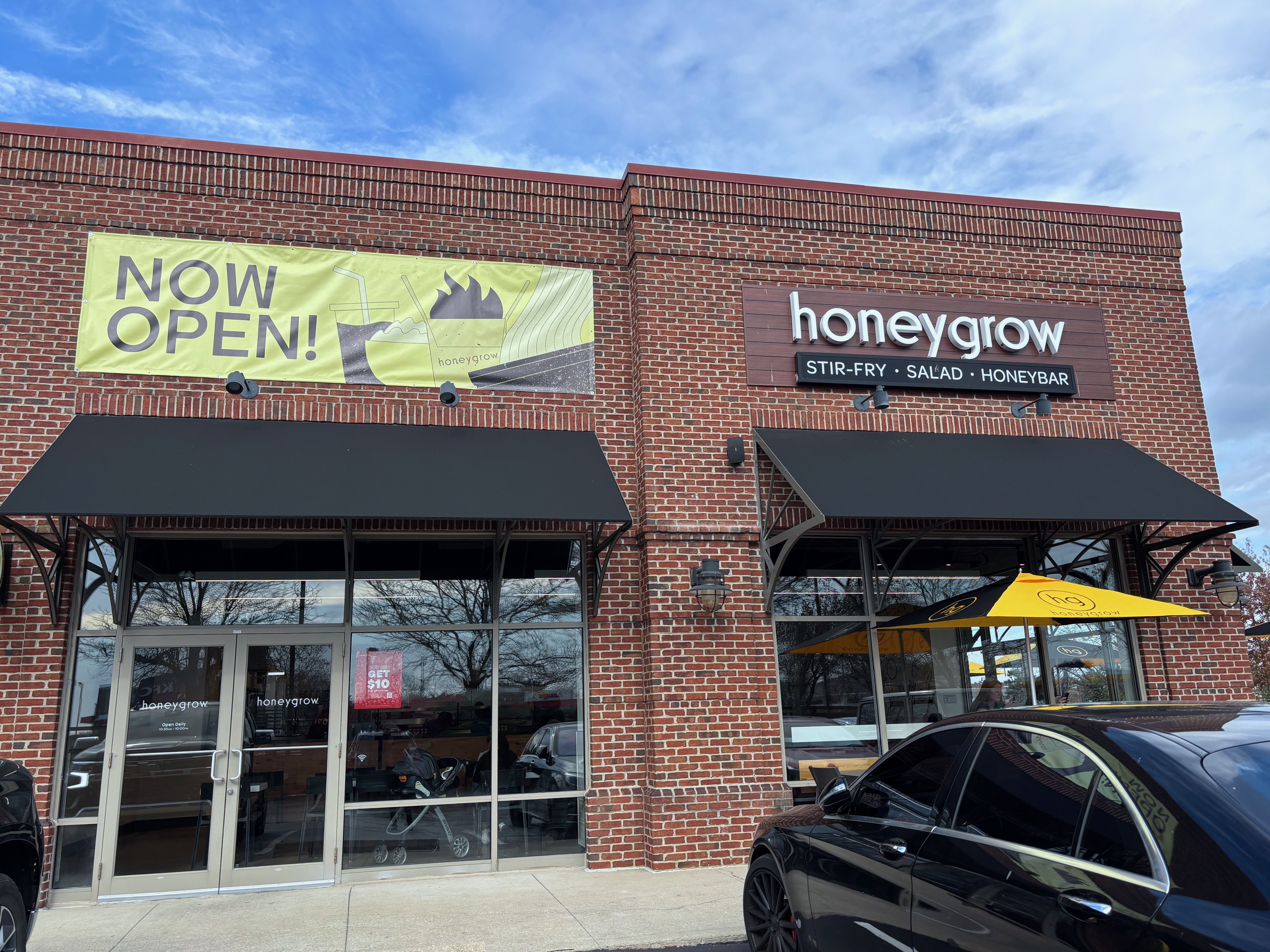 Brick storefront of honeygrow restaurant with black awnings, a black car parked outside, a yellow "Now Open!" banner, and a yellow honeygrow patio umbrella under partly cloudy sky.