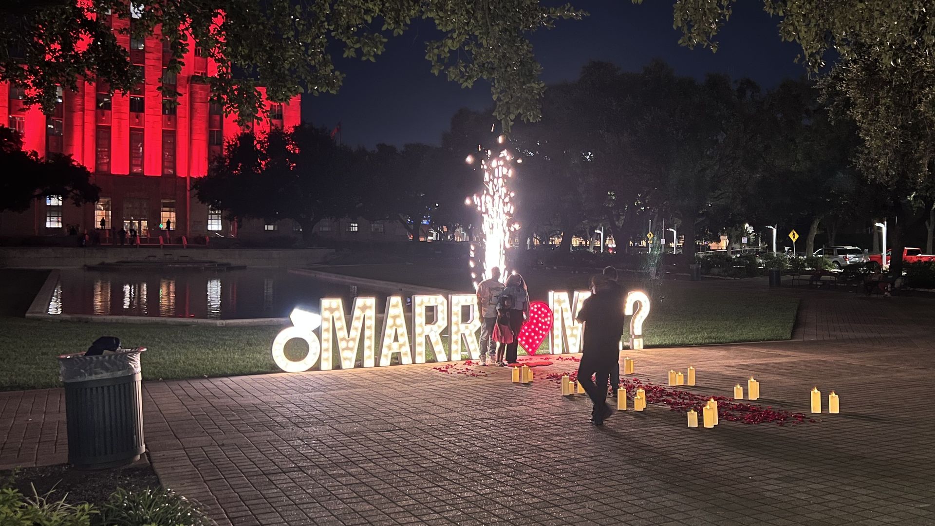A newly engaged couple stands in front of a sign reading "MARRY ME?" 