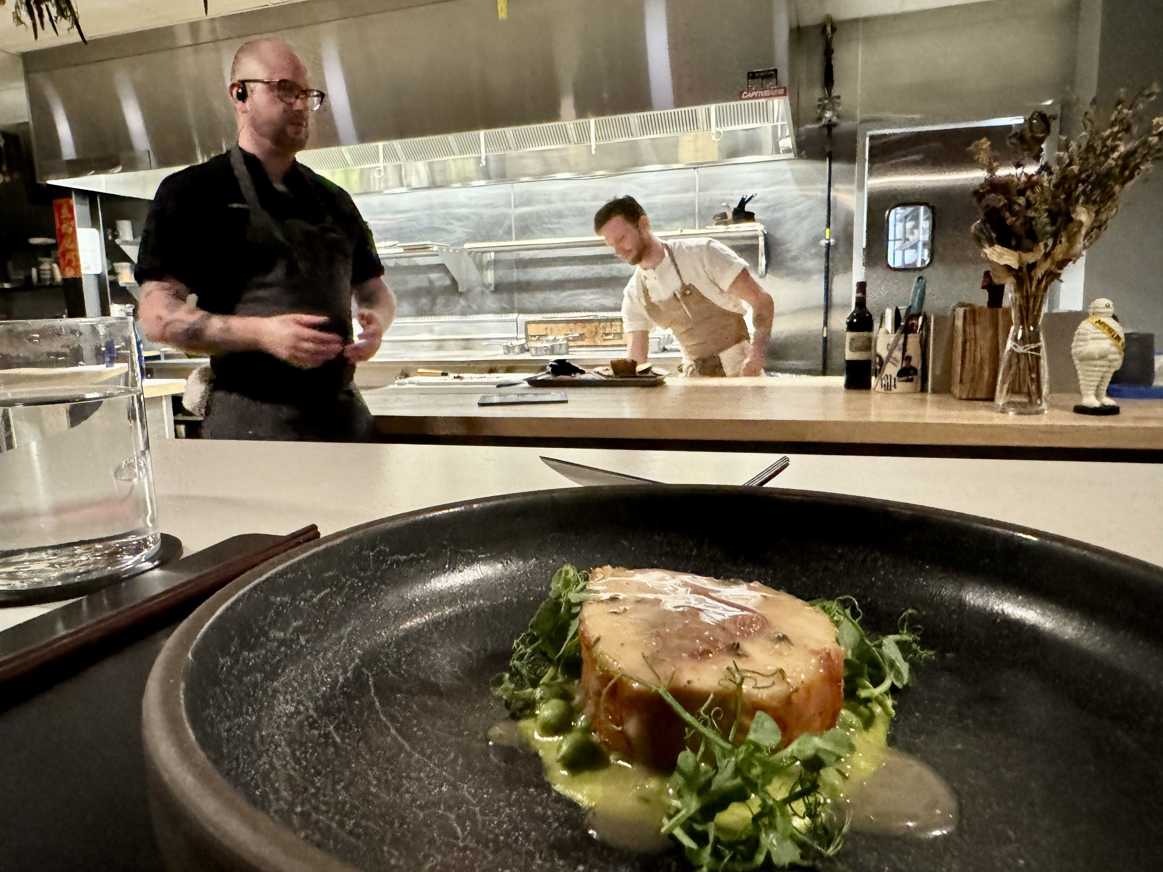 A plated dish of gourmet food on a restaurant counter, featuring a piece of meat surrounded by a green sauce and leafy greens. Two chefs are visible in the background in a professional kitchen. 